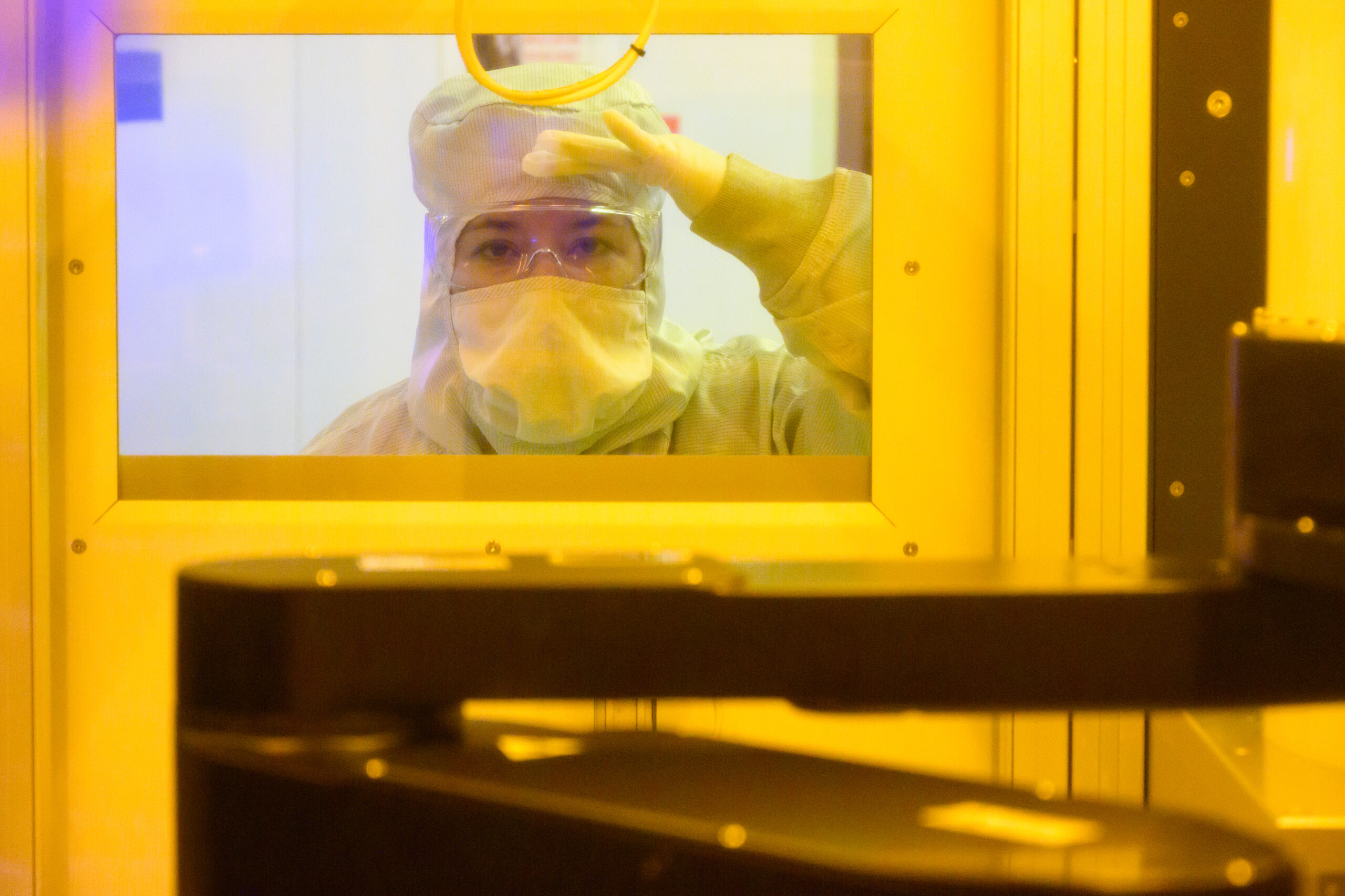 A person in protective cleanroom attire looks through a window, resting their hand on their forehead. The surrounding environment is dominated by a yellow hue, with industrial equipment in the foreground.