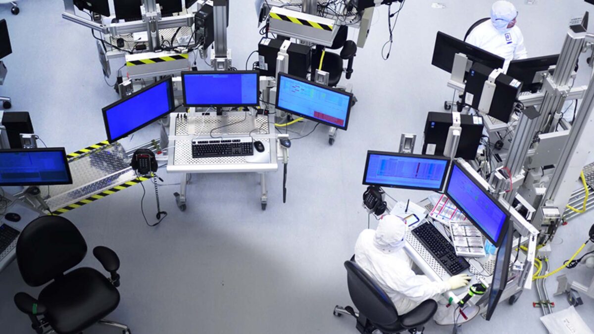 A high-tech laboratory with several workstations equipped with multiple monitors and computers. People in white protective suits are seated, working on the computers, surrounded by industrial equipment and cables on a clean, gray floor.