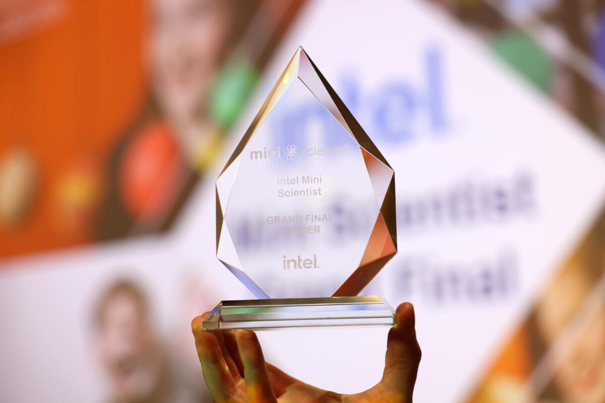 A hand holds a crystal award with the text Intel Mini Scientist Grand Final Winner engraved on it. The background is blurred, displaying colorful shapes and partial text.