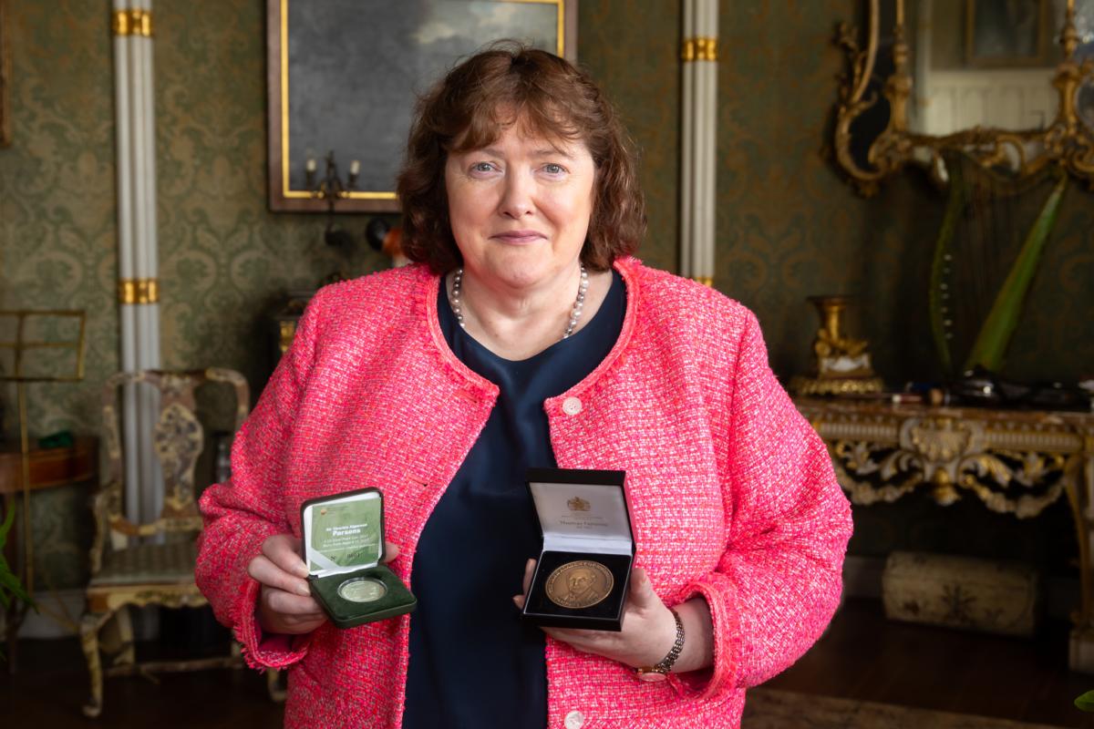 A person with brown hair wearing a pink jacket holds two open medal cases in a richly decorated room with ornate furniture and patterned wallpaper.