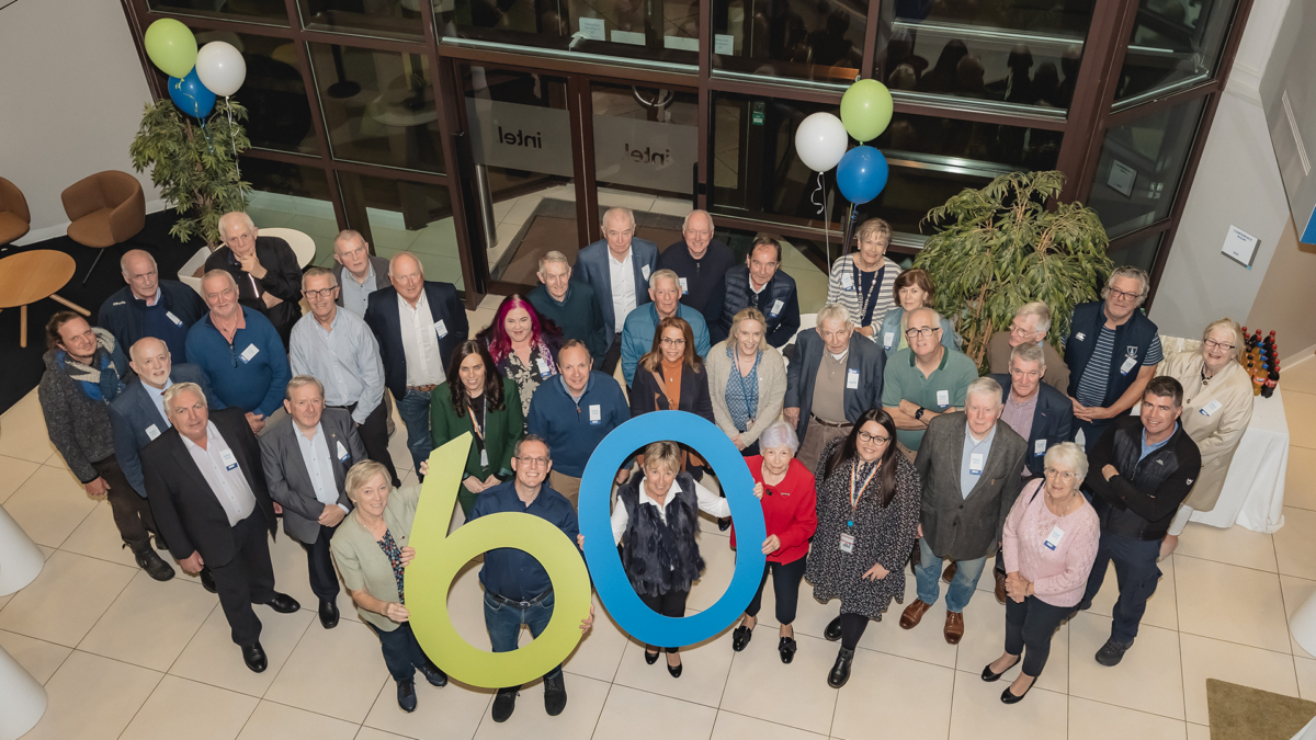 A group of people are gathered in a spacious room, holding large green and blue numbers 60 to celebrate an event. Some are wearing name tags. Balloons and large plants are in the background.