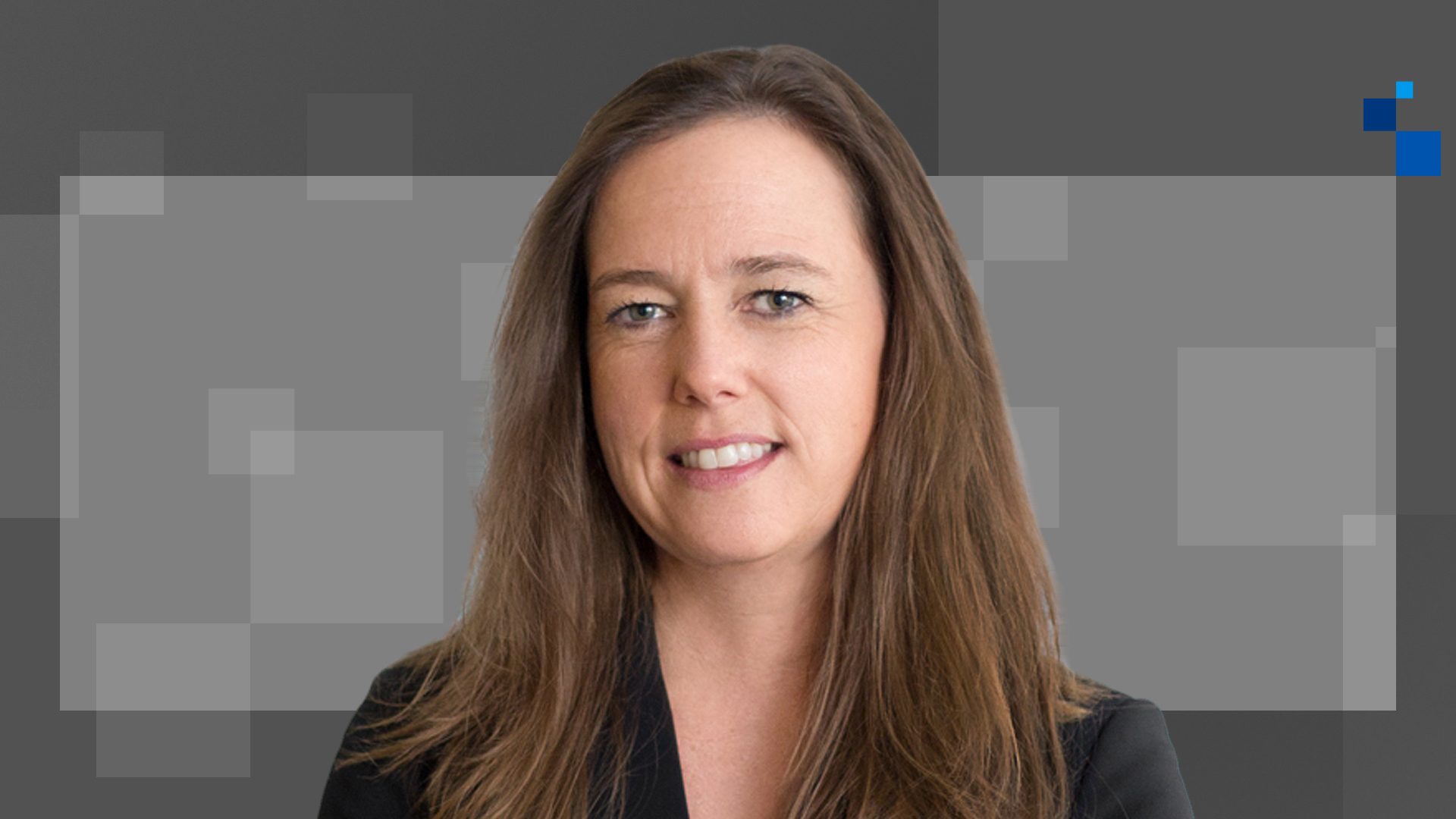 A woman with long brown hair is smiling against a geometric background with varying shades of gray. She is wearing a black jacket.