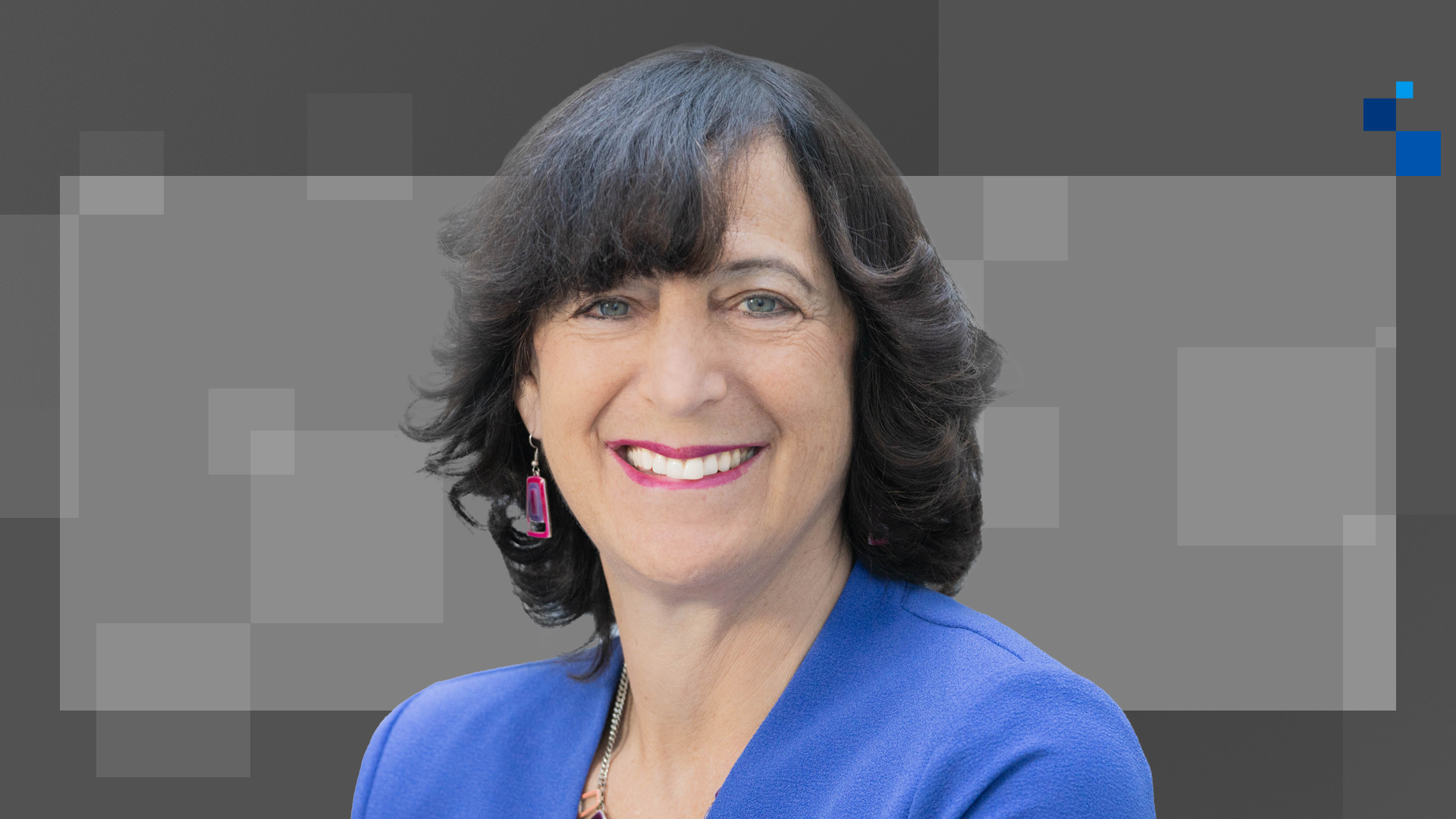 A person with shoulder-length dark hair and a bright smile is wearing a blue blazer and pink earrings. The background is a mix of light and dark gray squares.