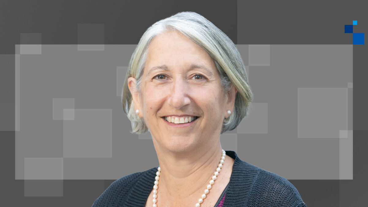 A smiling person with short gray hair wearing a black top and pearl necklace is in front of a gray patterned background with a blue square in the corner.