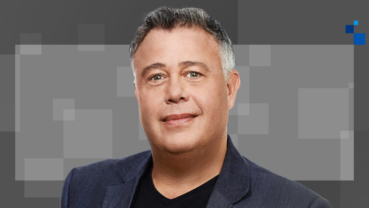 A person with short, graying hair is smiling softly at the camera. They are wearing a dark blazer over a black shirt. The background is a digital pattern of gray squares in varying shades.