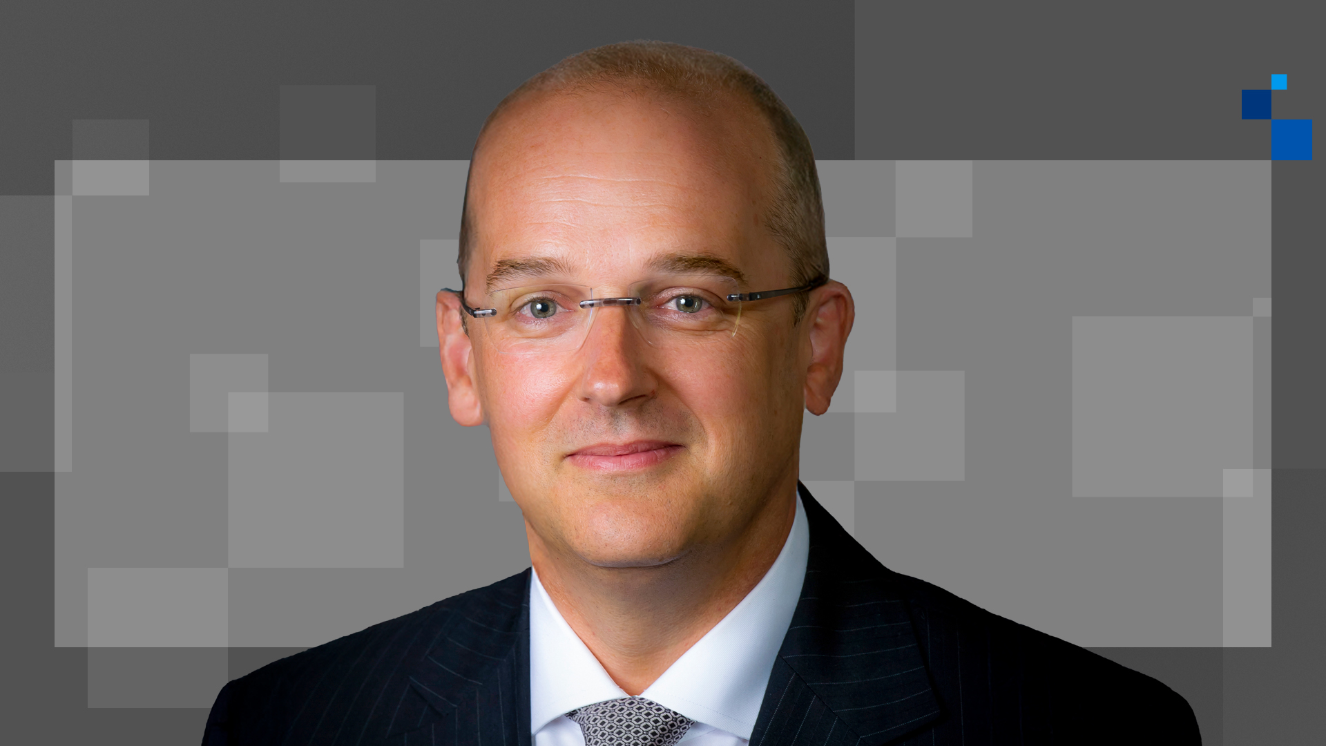 A man in a striped suit and tie, with glasses and short hair, is smiling slightly against a geometric background with gray squares.