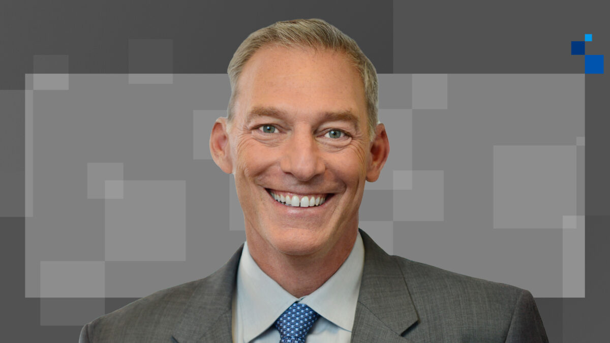 Smiling man in a gray suit and blue tie against a gray geometric background.