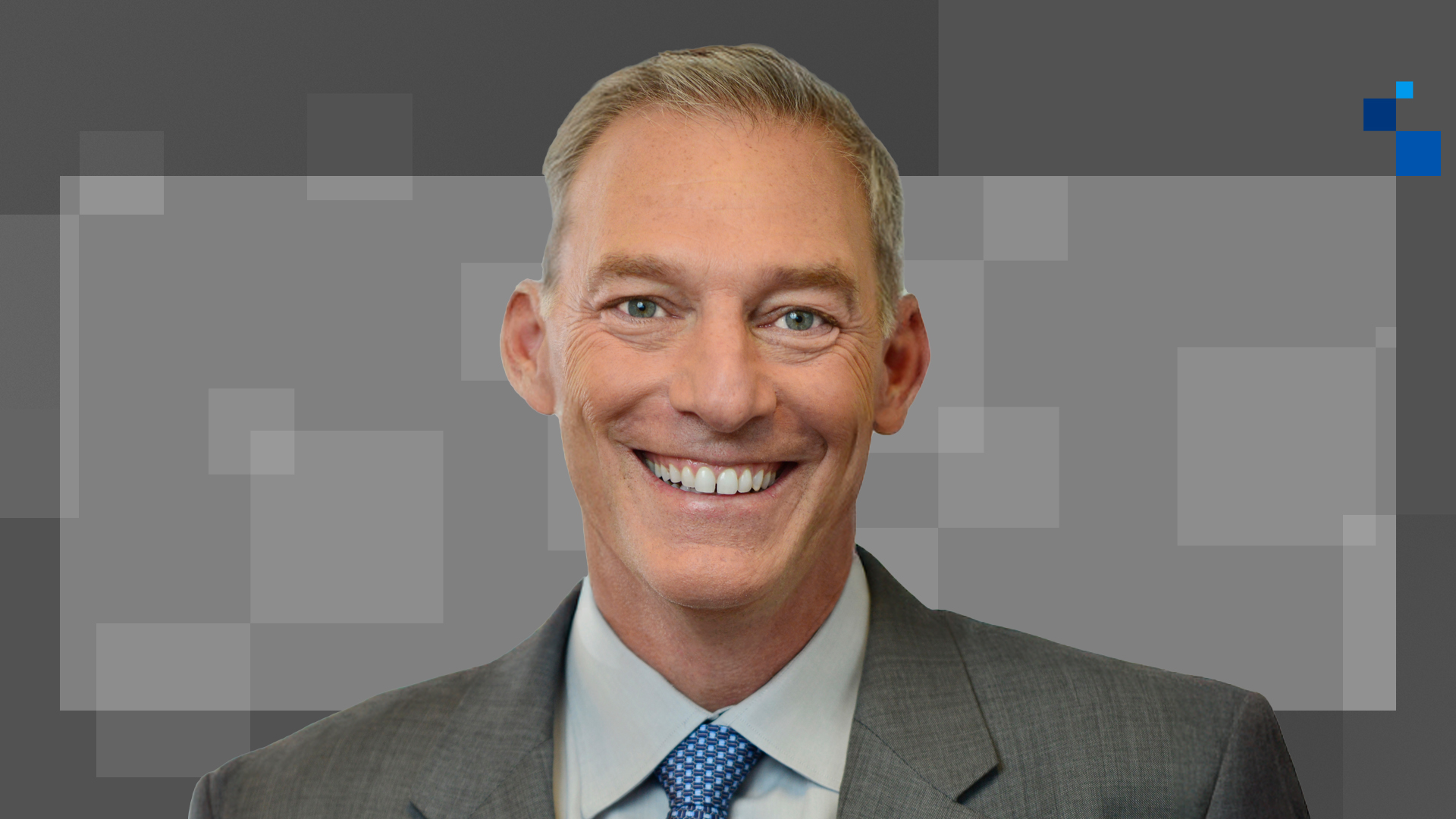 Smiling man in a gray suit and blue tie against a gray geometric background.