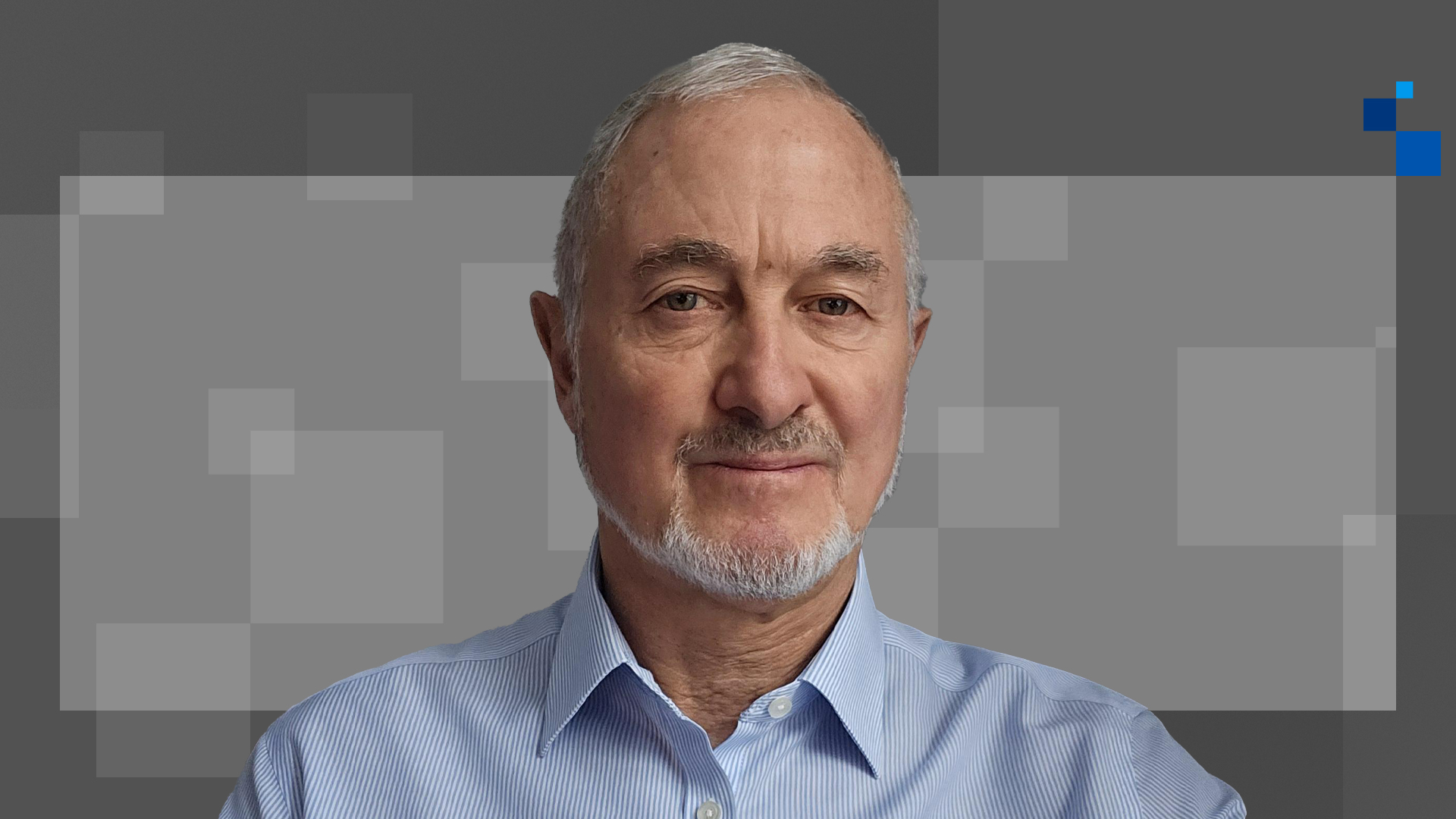 An older man with short gray hair and a beard wearing a light blue collared shirt stands in front of a gray background with square patterns.