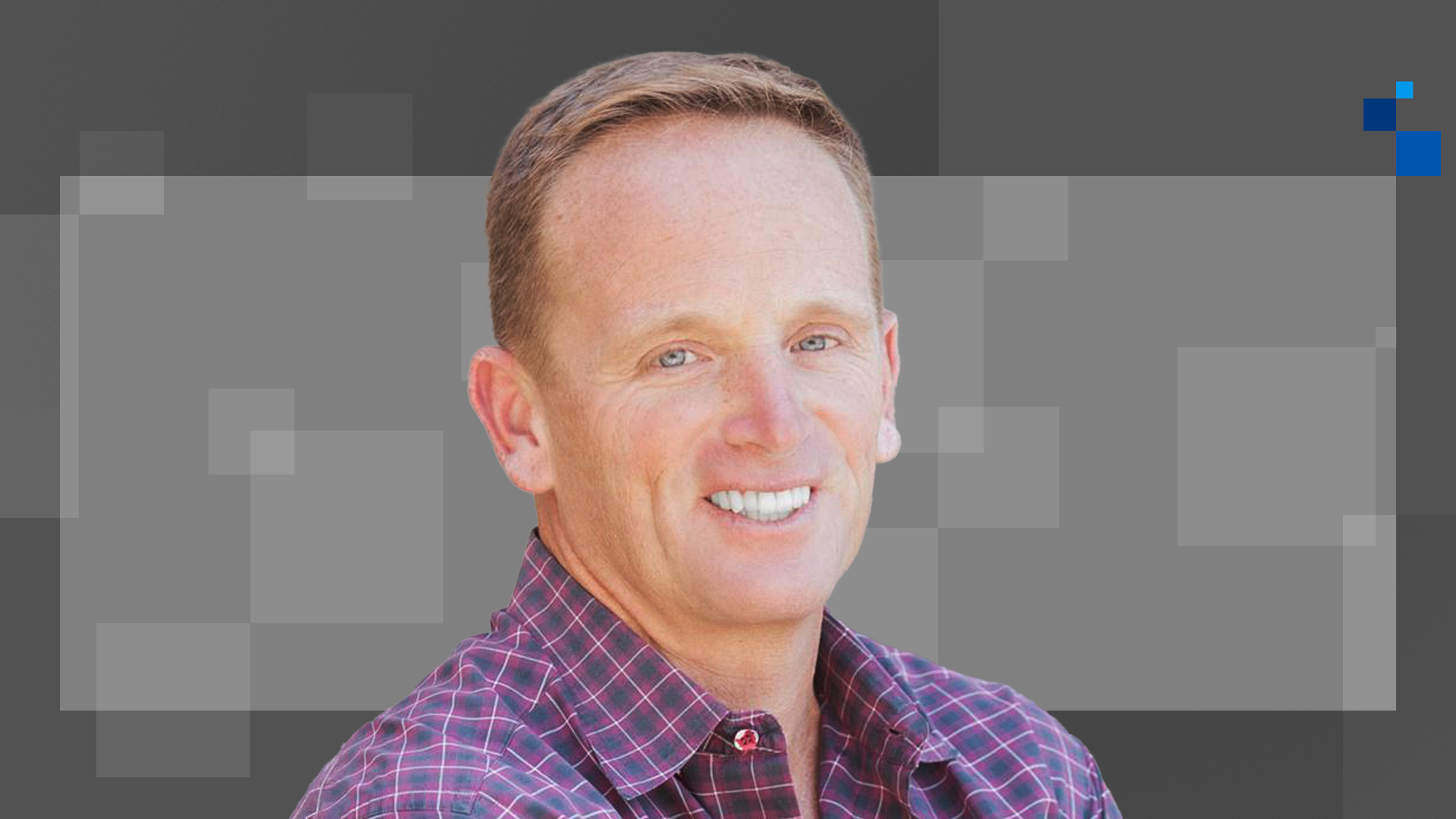 A person with short, light brown hair is smiling while wearing a checkered purple shirt against a gray, pixelated background.