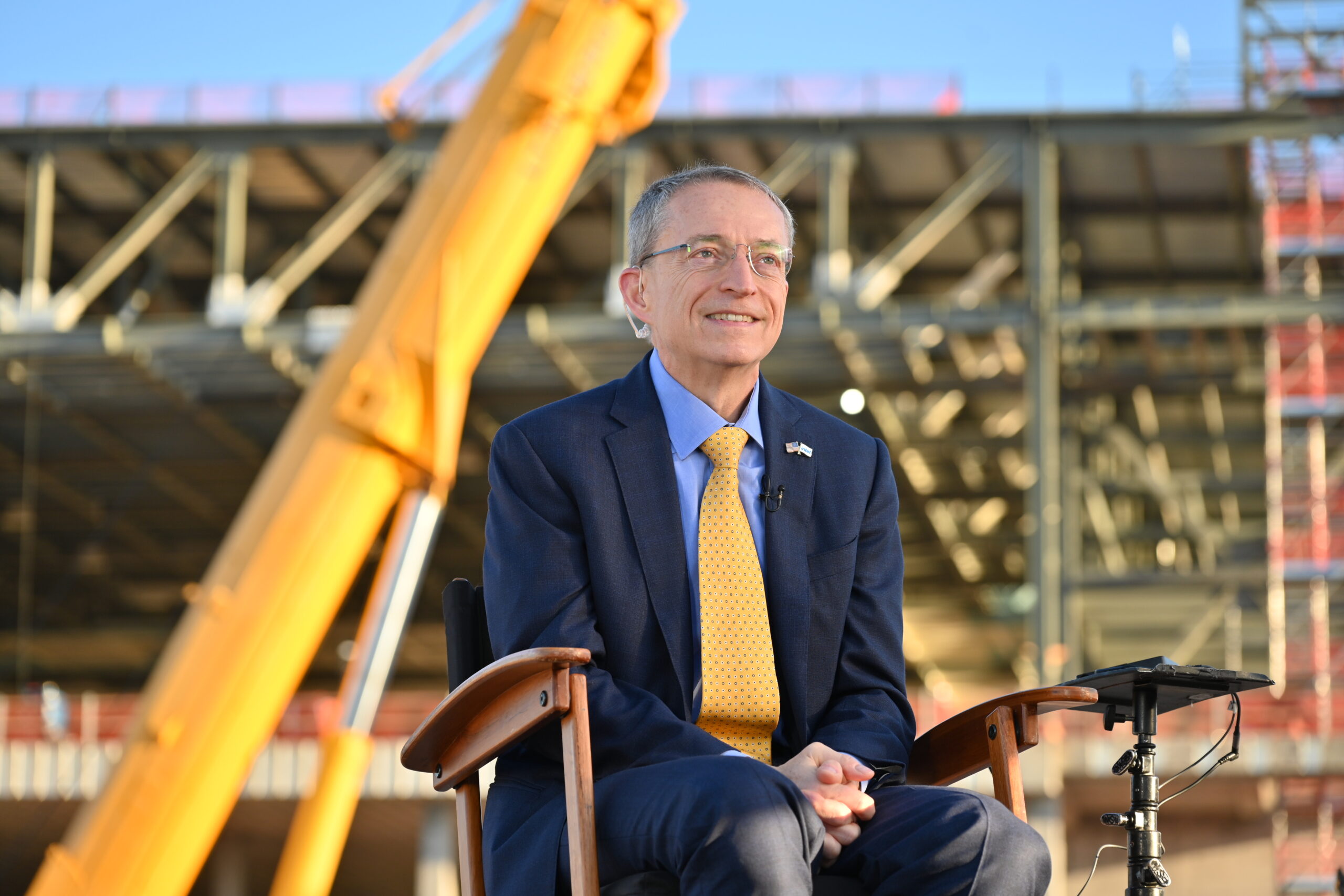 A person in a suit and yellow tie is seated outdoors at a construction site. A large crane is in the background, along with steel framework of a building under construction. The individual is smiling and there is a microphone stand nearby.