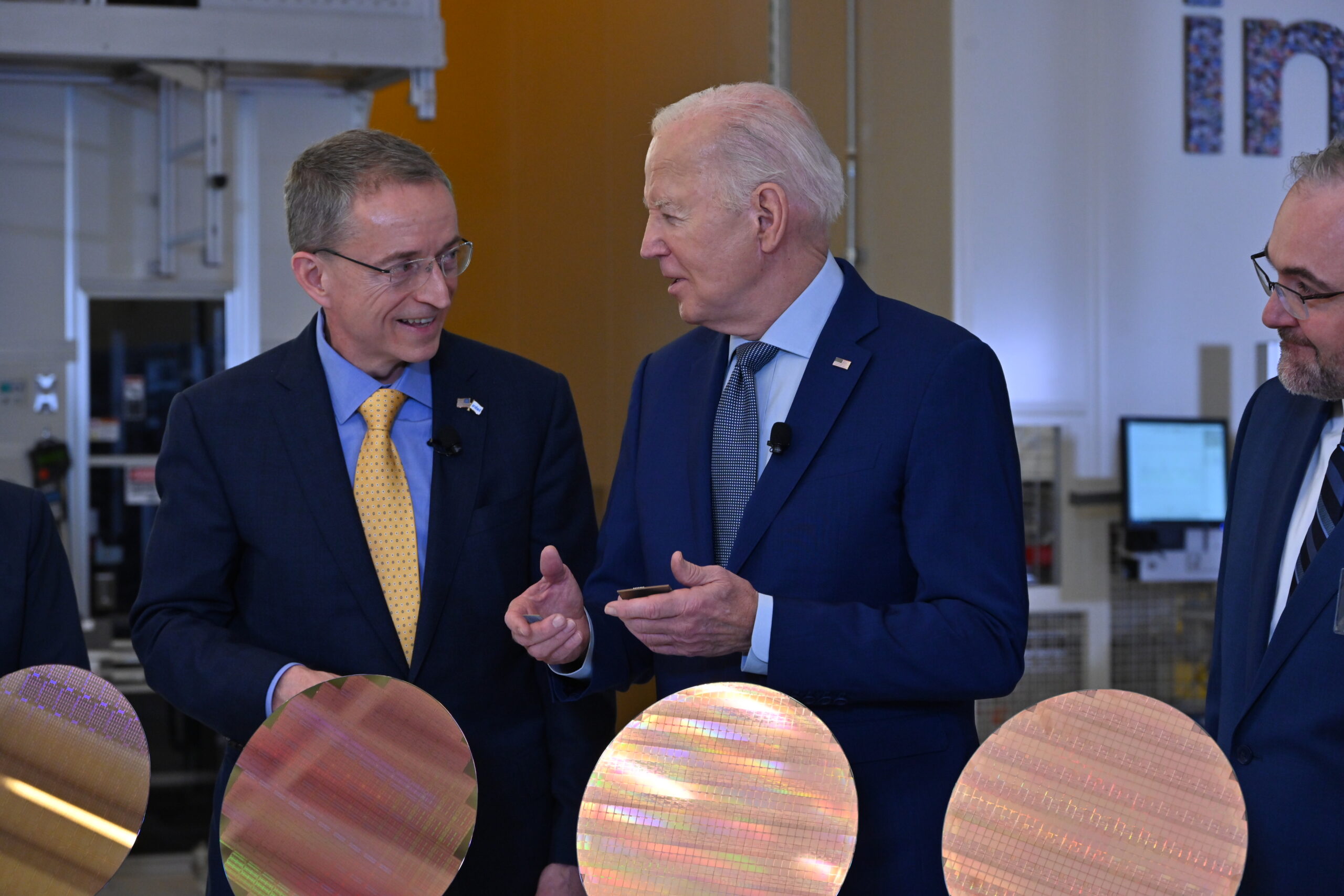 Three men in suits stand in a technology facility, engaged in conversation. One in the center gestures with his hands. In front of them are several large, round silicon wafers. They appear to be discussing technology or manufacturing.