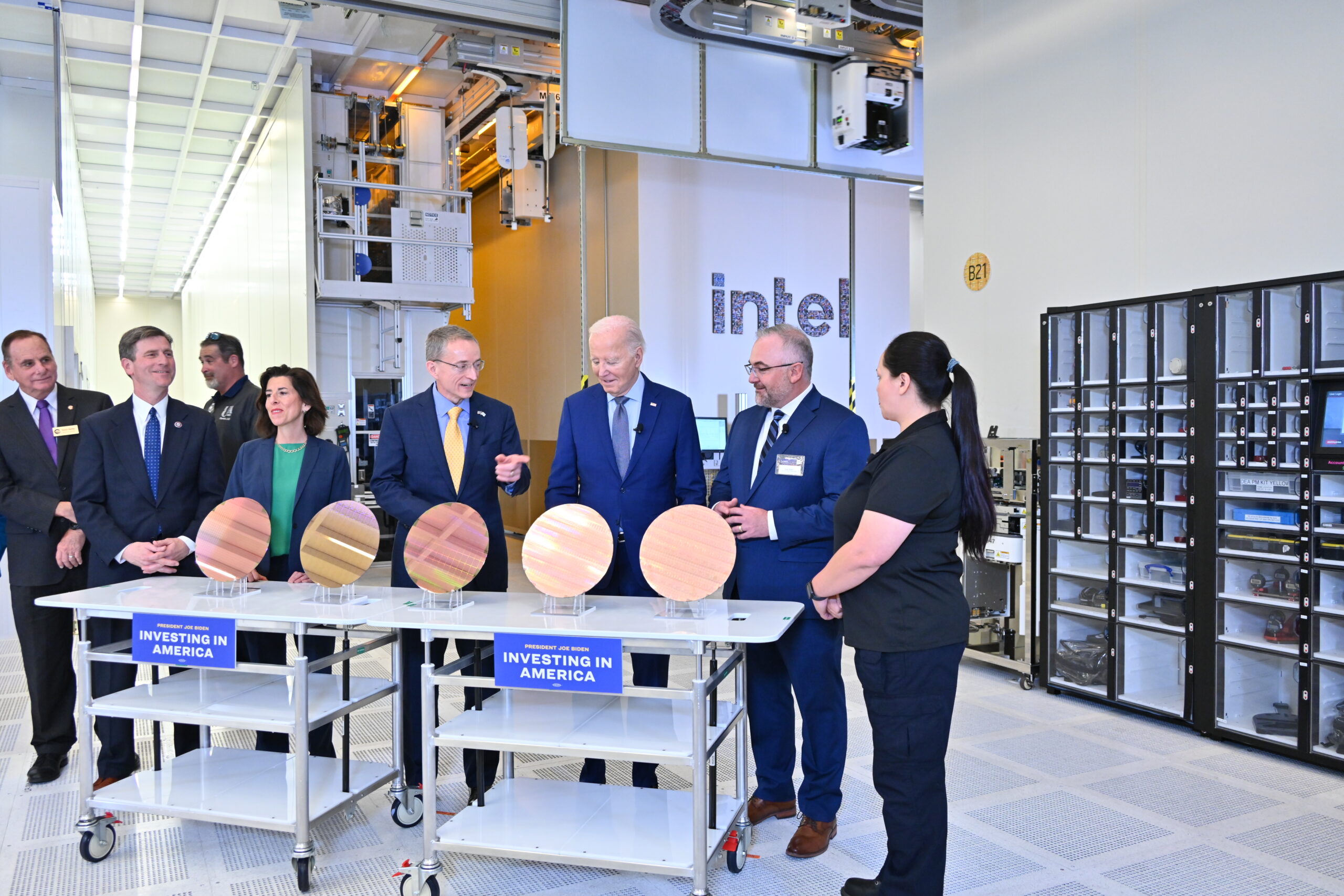 A group of people in formal attire stands in a high-tech facility with Intel in the background. They are gathered around tables displaying silicon wafers. Signs on the tables read Investing in America. The room is filled with advanced equipment.