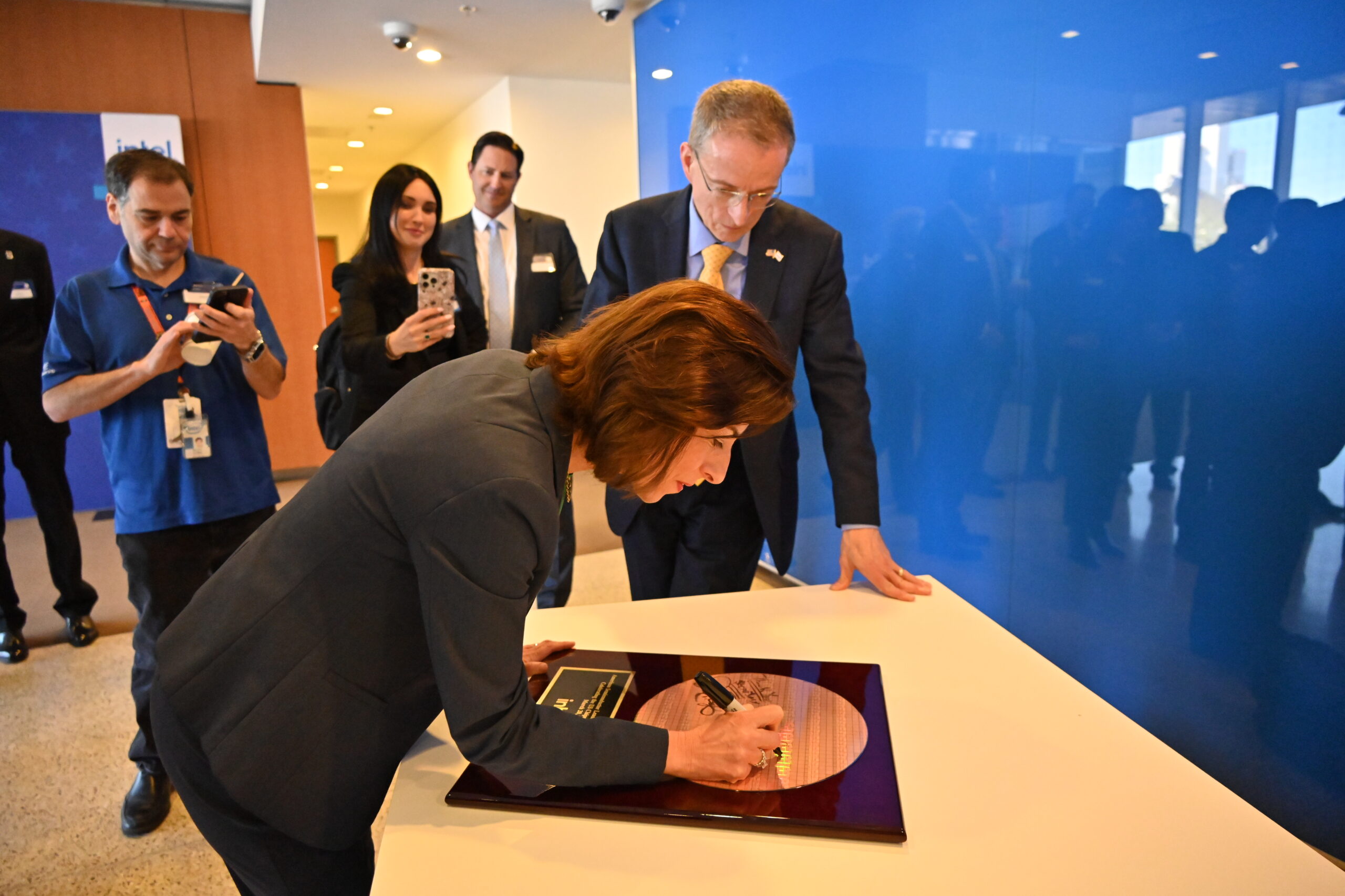 A woman signs a large wafer mounted on a plaque at a ceremony, while a man in a suit watches. Several people stand in the background, some taking photos. The setting appears to be a professional or corporate environment.