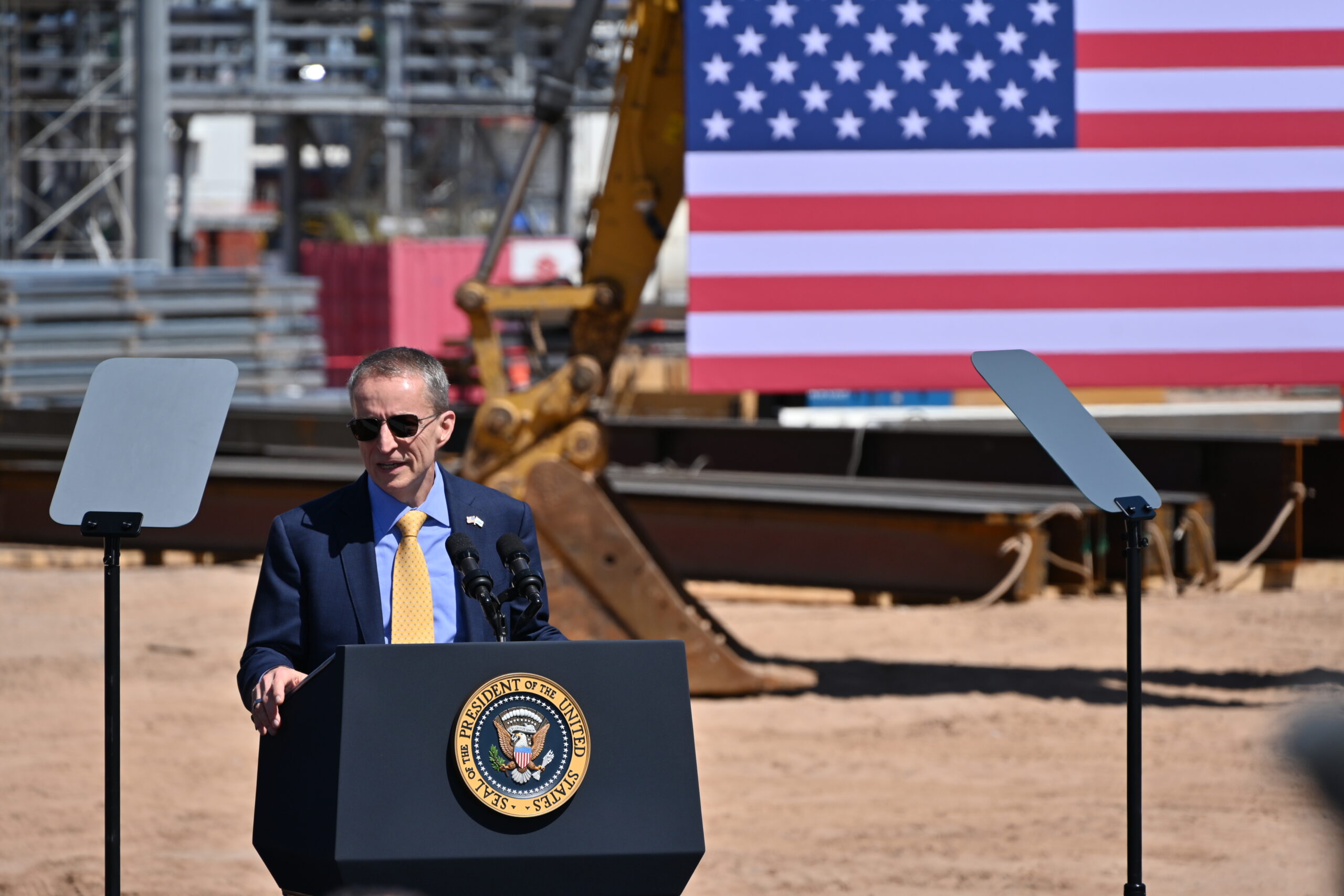 A man in a suit and sunglasses speaks at a podium with the presidential seal. Two teleprompters stand nearby. Behind him, a large American flag and construction equipment are visible under a clear blue sky.