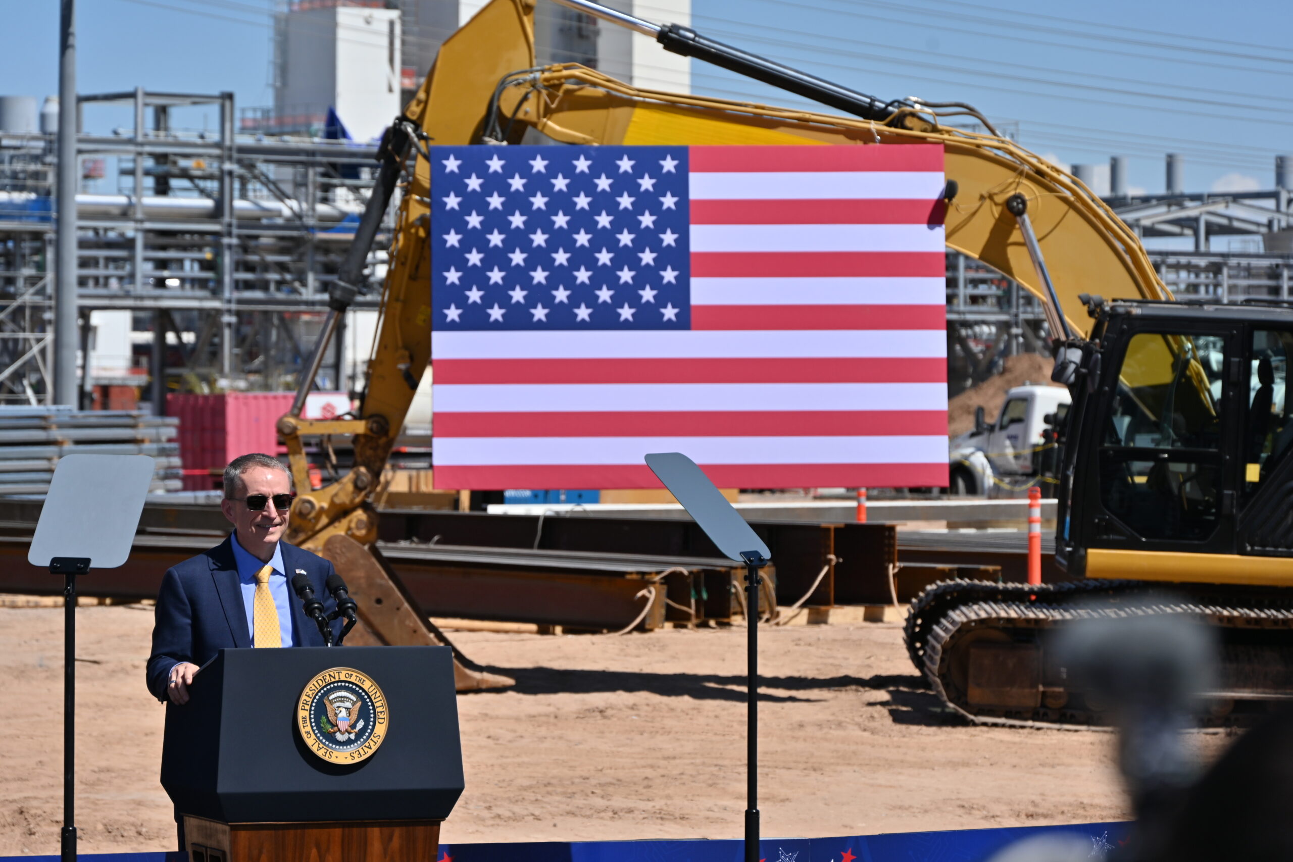 A man stands at a podium with the U.S. presidential seal, speaking into microphones. Behind him, a large American flag is displayed on a construction site with an excavator. Industrial structures are visible in the background.