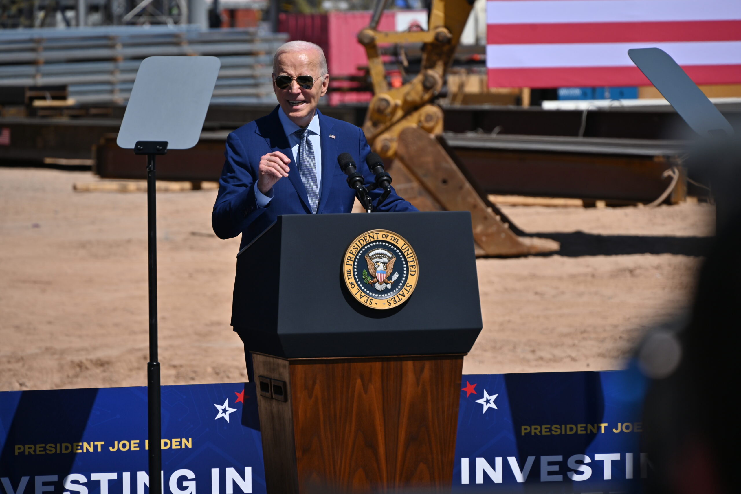 A person in a suit speaks at a podium with the presidential seal. The backdrop includes construction equipment and an American flag. Signs reading Investing in are partially visible.
