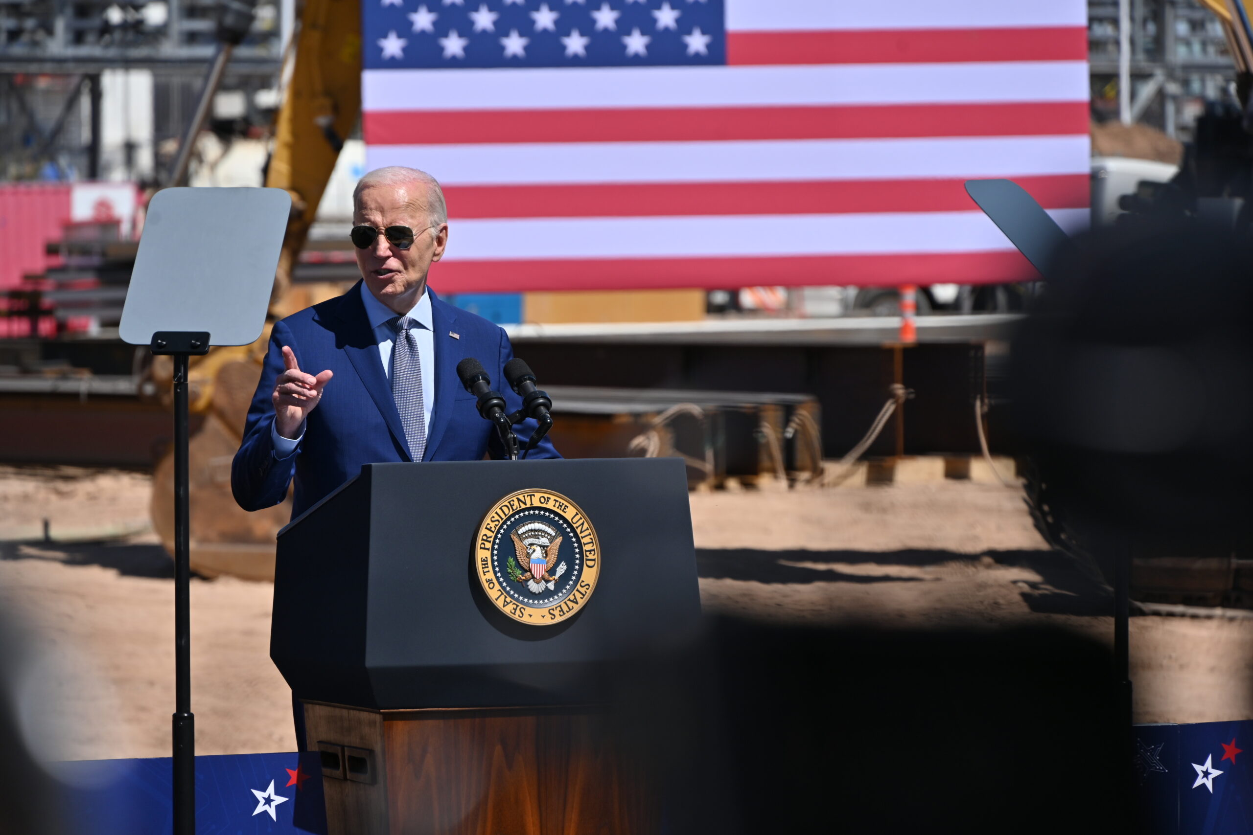 A man in a blue suit and sunglasses speaks at a podium with the presidential seal. An American flag is displayed in the background. Construction equipment is visible, suggesting a worksite setting.