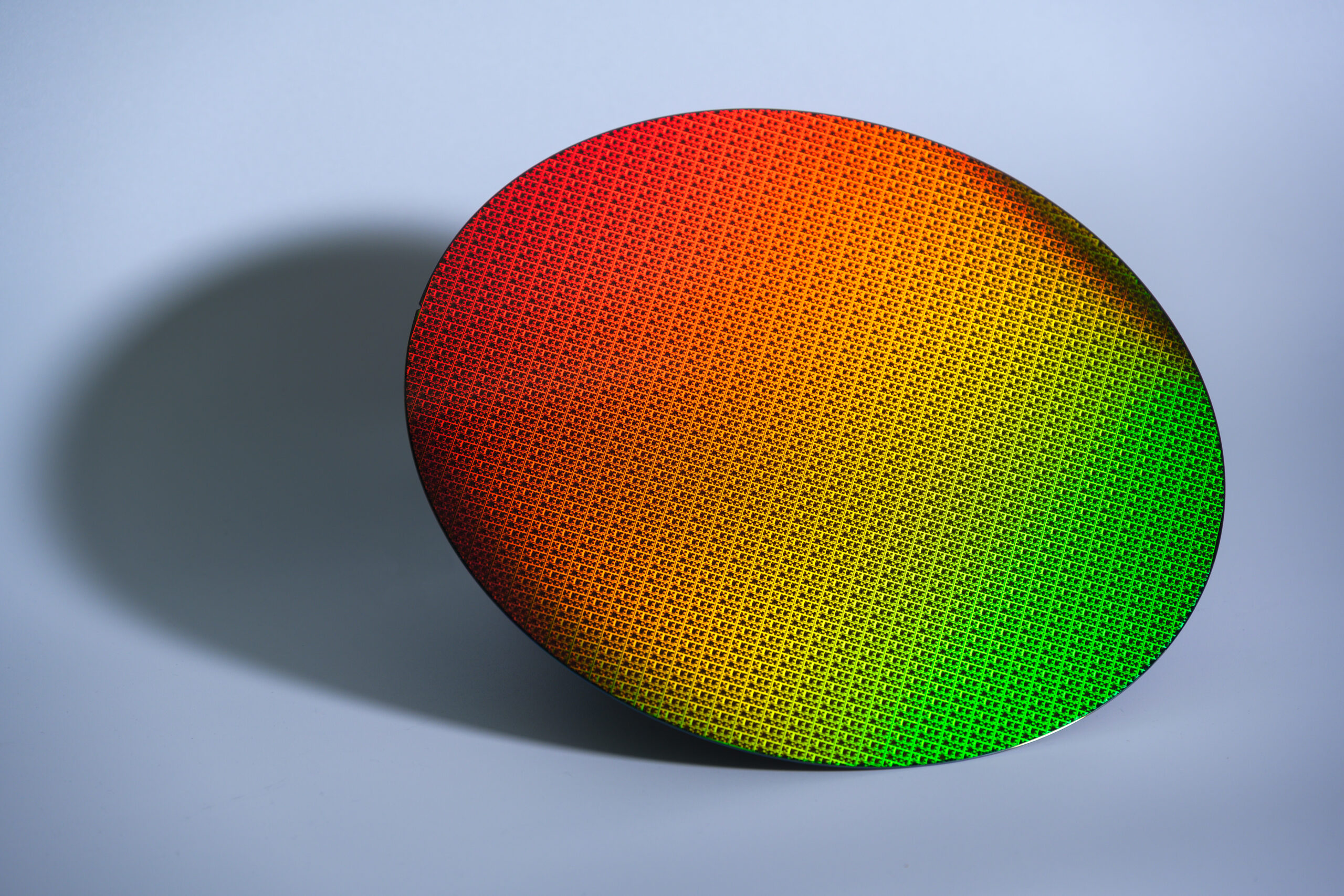A colorful silicon wafer sits against a light blue background, showcasing a gradient pattern transitioning from red to green. The intricate, reflective surface displays microchip circuits, casting a subtle shadow to the left.