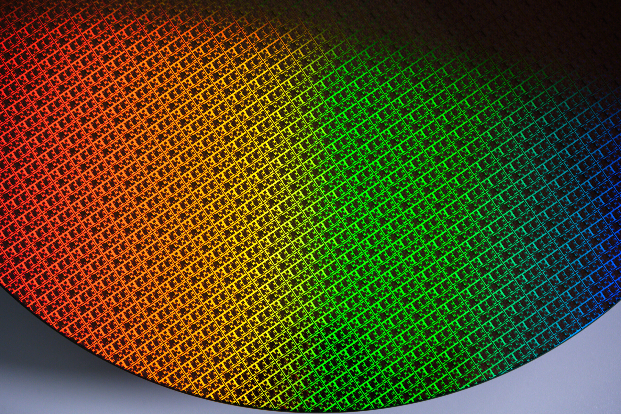 A close-up of a brightly colored silicon wafer, displaying a grid pattern. The surface reflects a spectrum of colors, blending from orange on the left to green on the right, showcasing the intricate and reflective nature of the wafer.