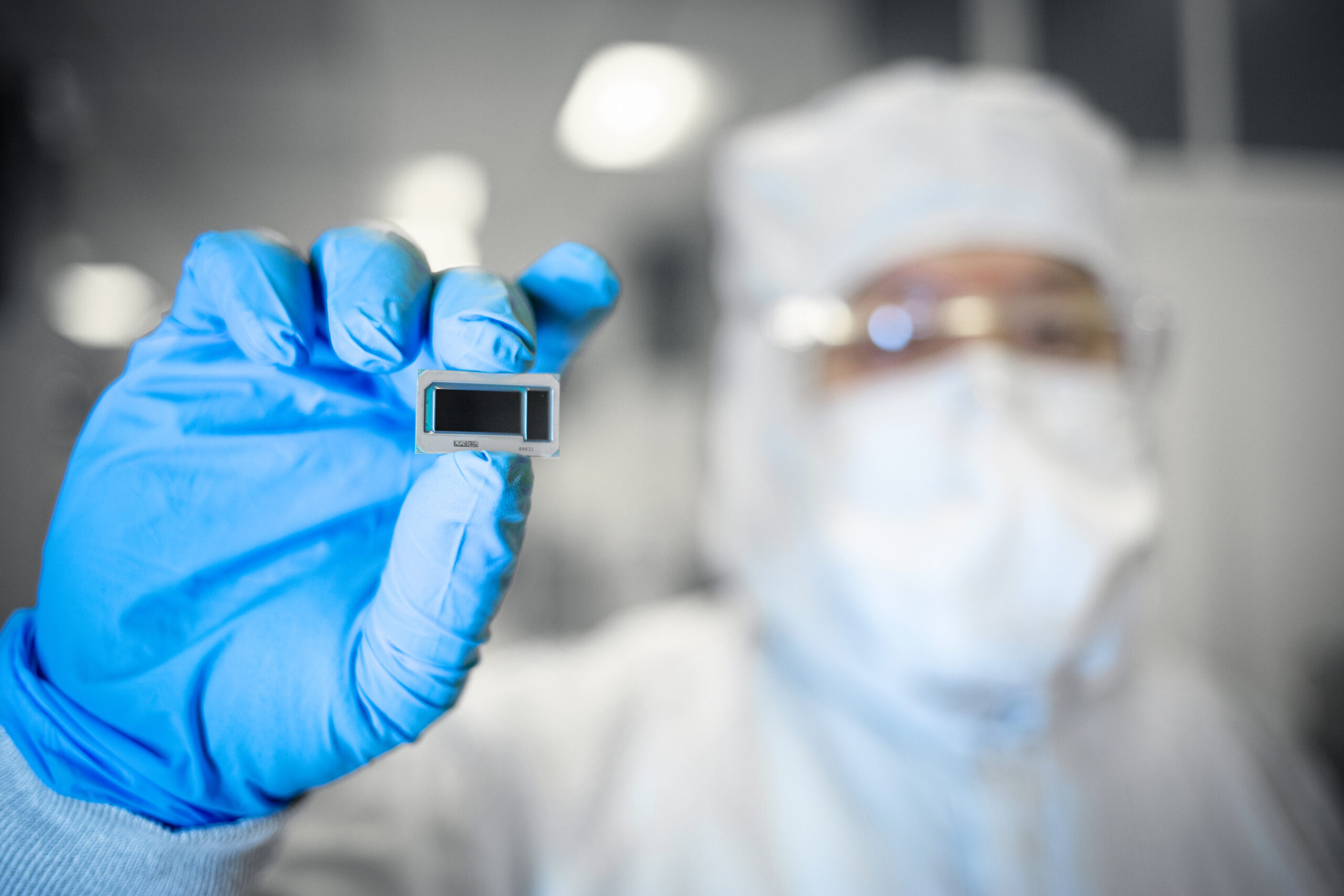 A person wearing protective clothing and gloves holds a small microchip in focus. The background is blurred, highlighting the microchips details. The individual is in a laboratory or clean room setting.