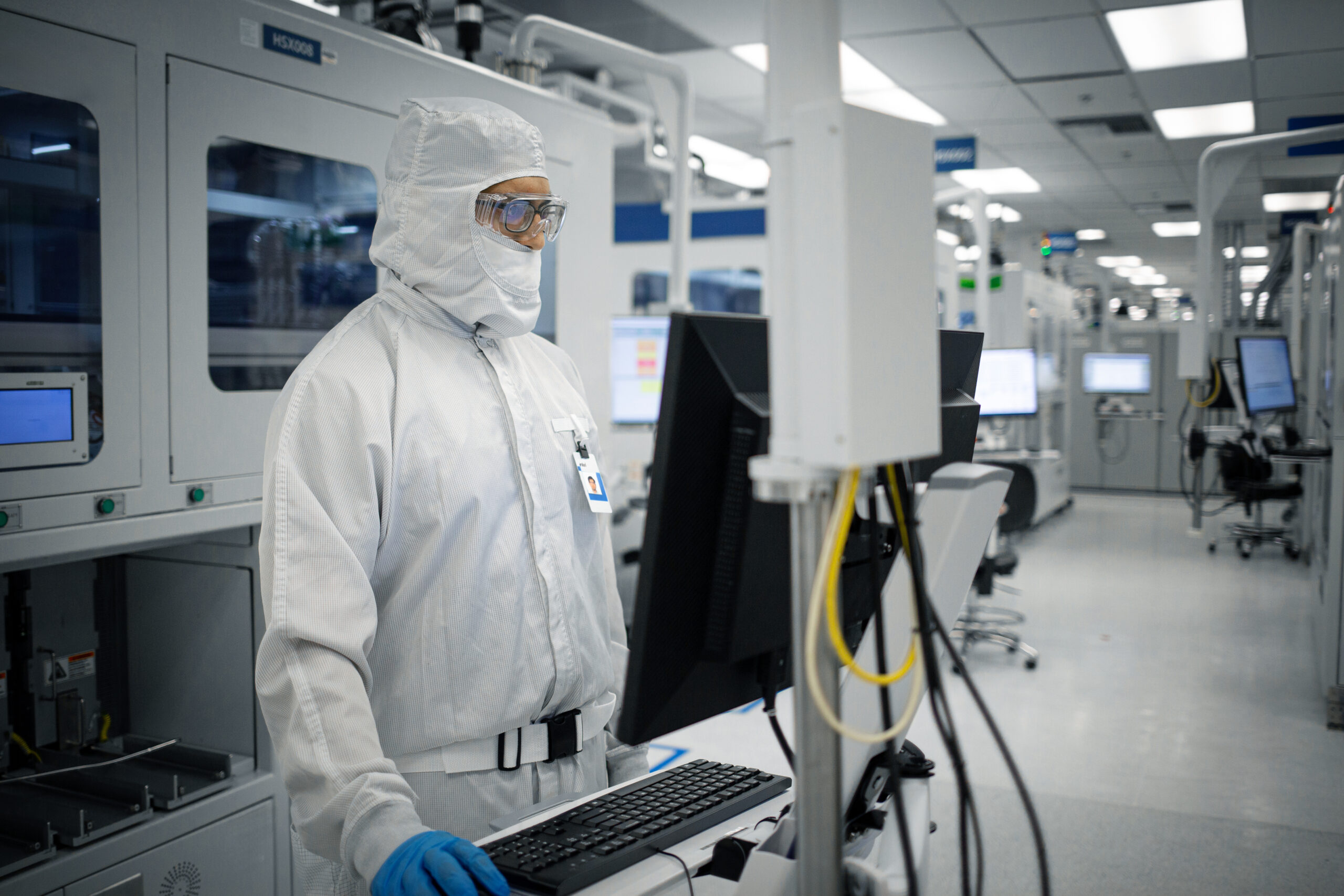 A person in a full protective suit, including a face mask and gloves, stands in a modern, clean laboratory environment focused on a computer screen. The setting features advanced machinery and bright overhead lighting.