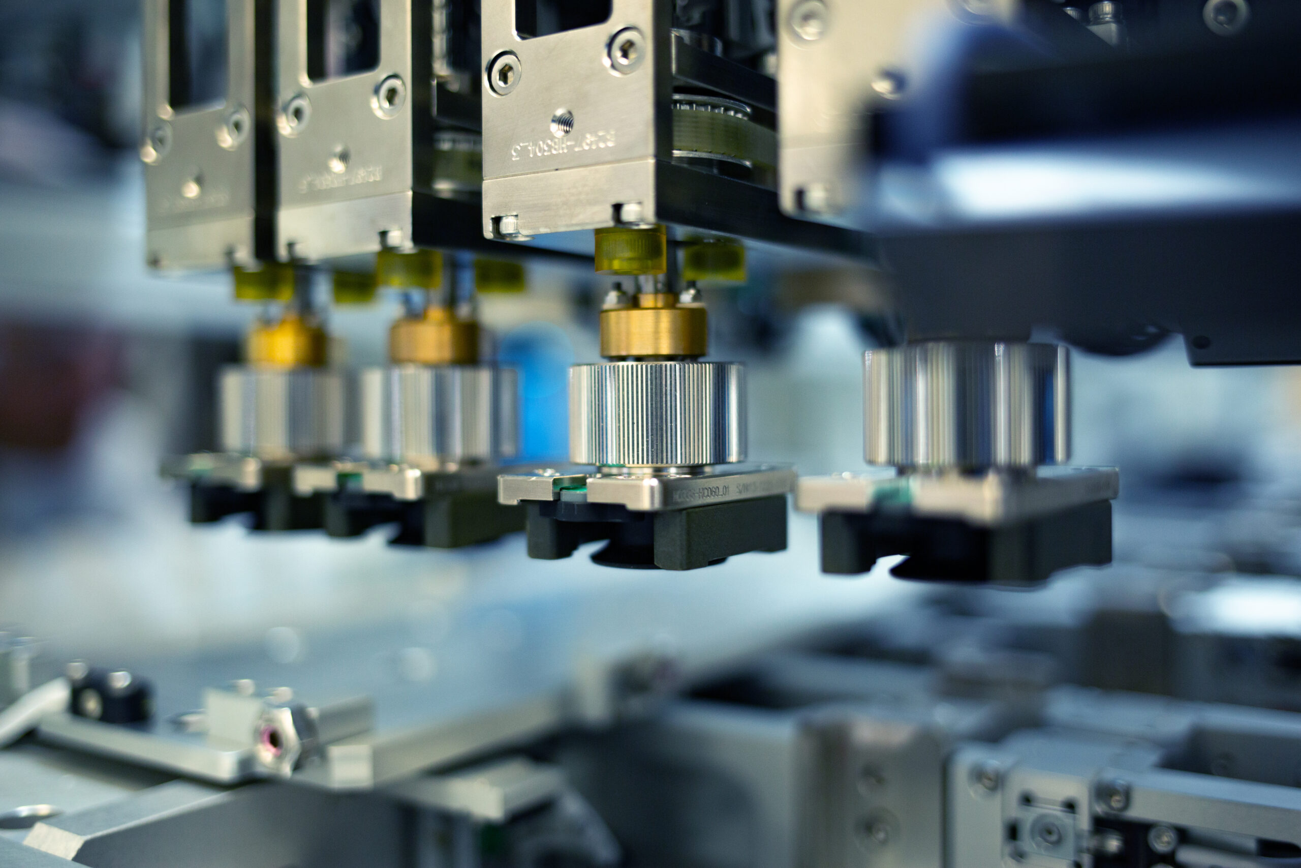 Close-up of robotic arms in a factory setting, each equipped with precision tools, positioned over a metallic surface. The background is slightly blurred, emphasizing the focus on the machinerys intricate components.