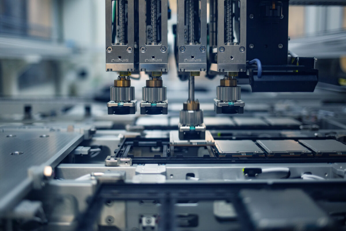 A close-up view of an industrial machine with multiple robotic arms working on an assembly line in a factory setting. The machinery is equipped with various components, indicating high-tech manufacturing or electronics production.
