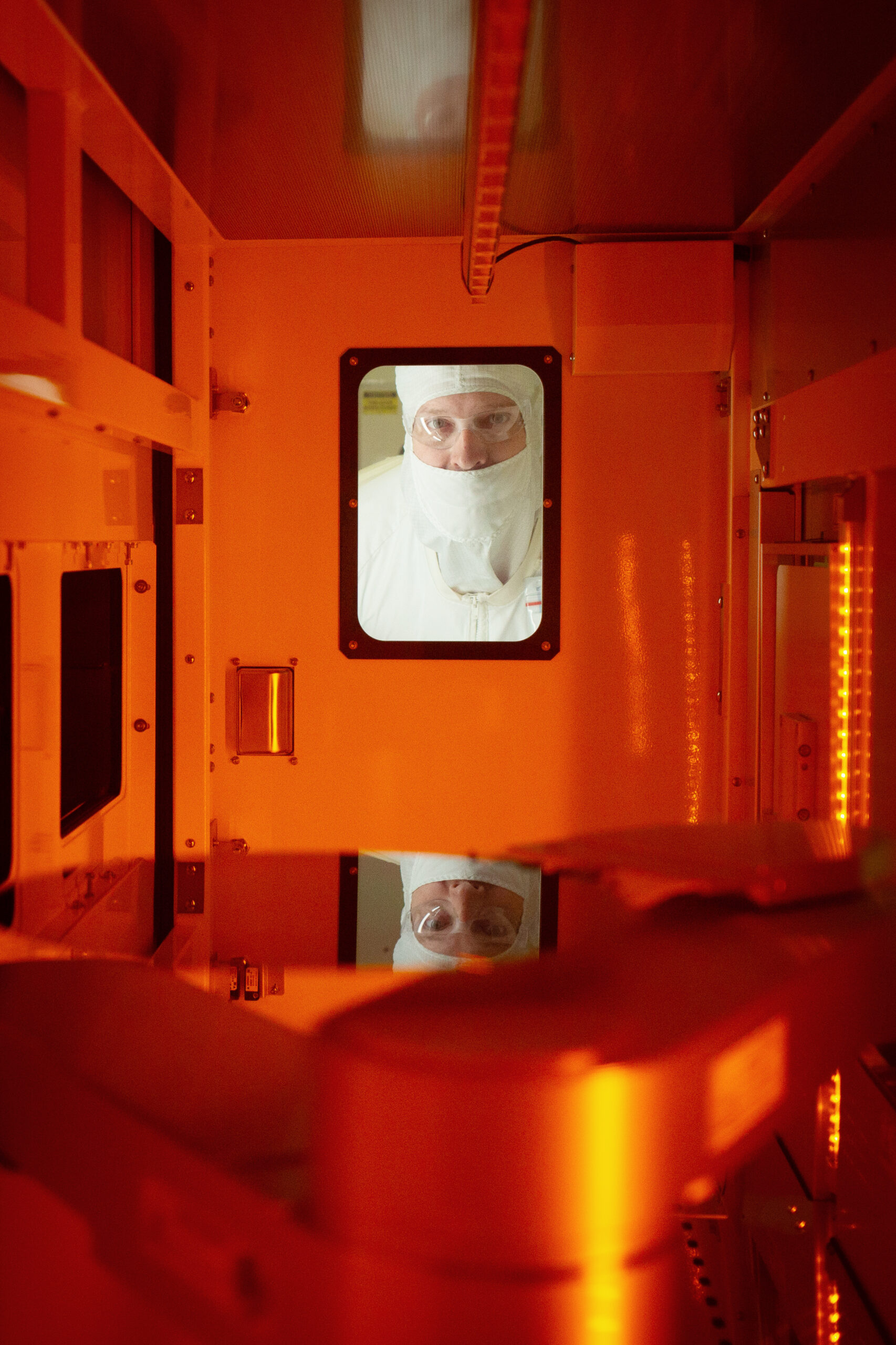 A person wearing a protective suit and mask is looking through a small window in a lab setting with orange lighting. The interior features reflective surfaces and machinery.