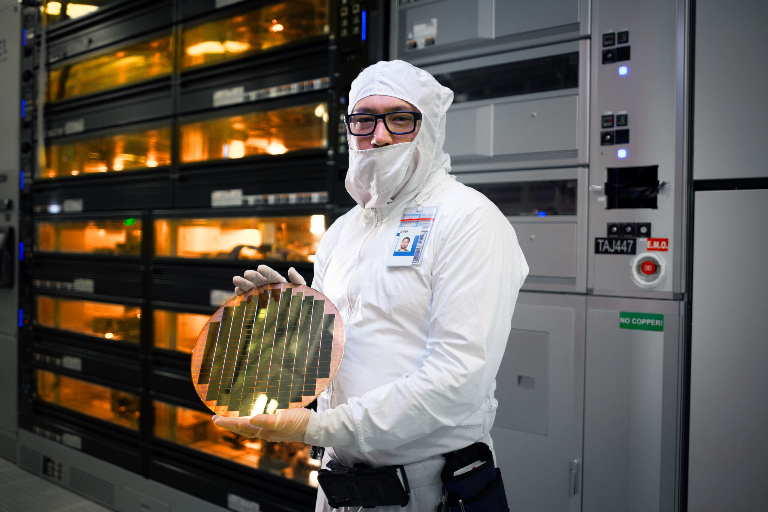 A person in a cleanroom suit holds a semiconductor wafer. They stand in a high-tech lab environment, surrounded by advanced equipment and large storage units. The atmosphere is clean and high-tech.