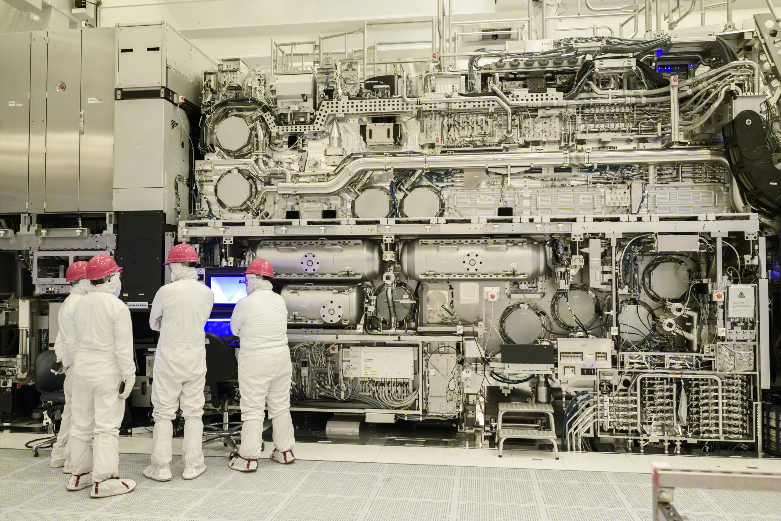 Four people in white protective suits and pink helmets stand in front of a large, complex machine with interconnected components and wiring, working on a computer. The setting appears to be a cleanroom environment in a technology facility.