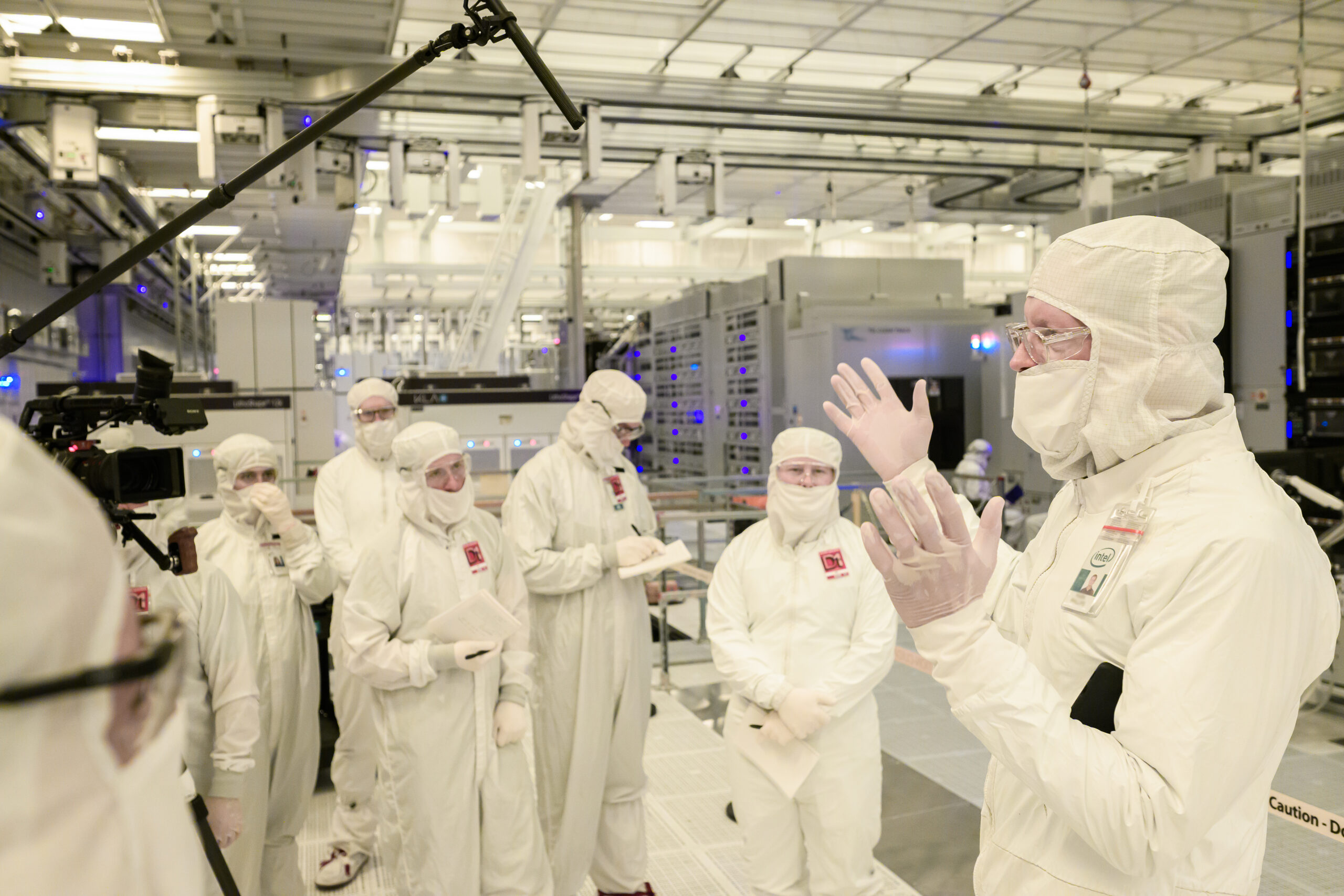 A group of people in white cleanroom suits and masks stand in a high-tech laboratory. One person gestures while speaking to the group. A camera on a boom captures the scene, and various technical equipment is visible in the background.