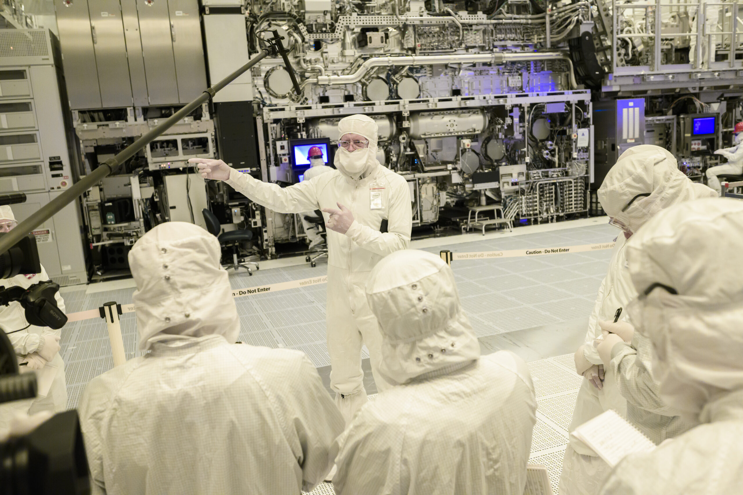 A group of people in cleanroom suits gather around a person who is speaking and gesturing inside a high-tech laboratory with complex machinery. The environment is sterile, with visible equipment and monitors in the background.