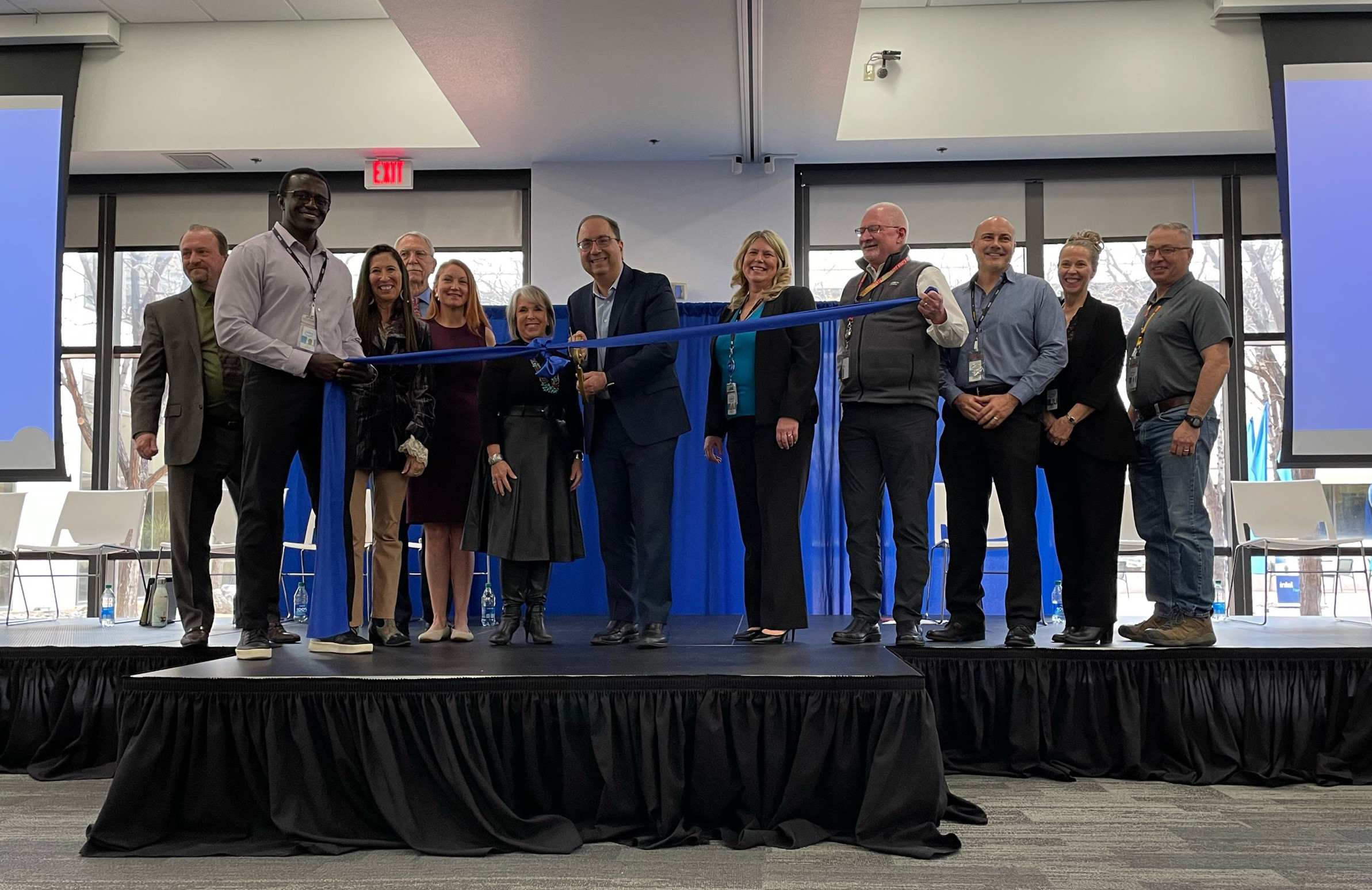 A group of people standing on a stage during a ribbon-cutting ceremony. One person is cutting the ribbon with large scissors. Everyone is smiling, and there are two large projection screens in the background.