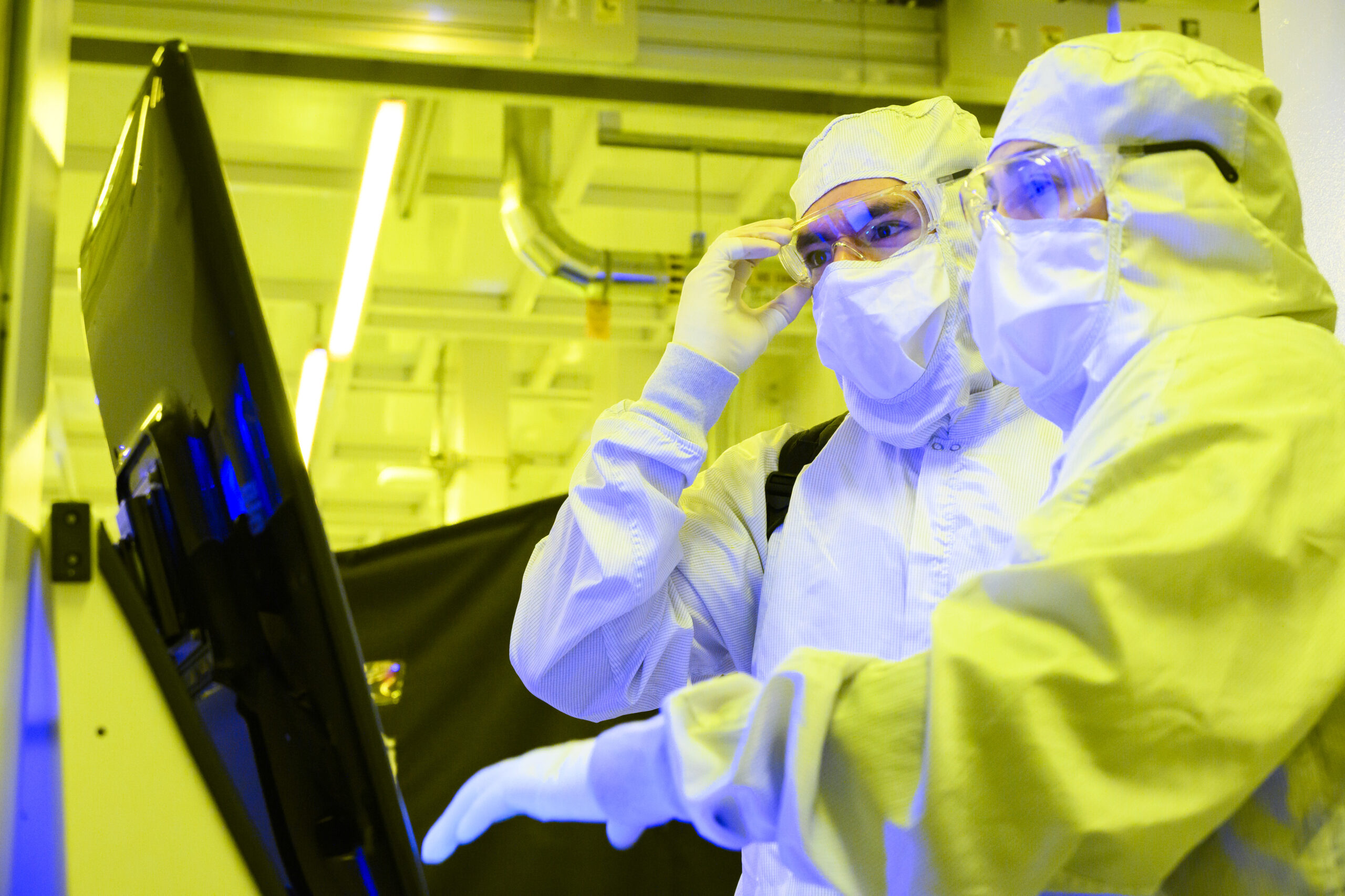 Two individuals in protective suits and goggles work in a lab illuminated with yellow light. They are focused on a large screen, likely engaged in a scientific or technical task.