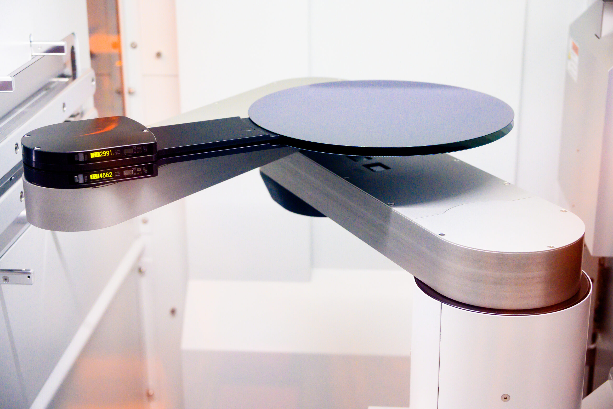 A robotic arm holds a silicon wafer in a semiconductor manufacturing facility. The setting is highly technical and sterile, with metallic surfaces and precision equipment, illustrating advanced technology and automation in action.