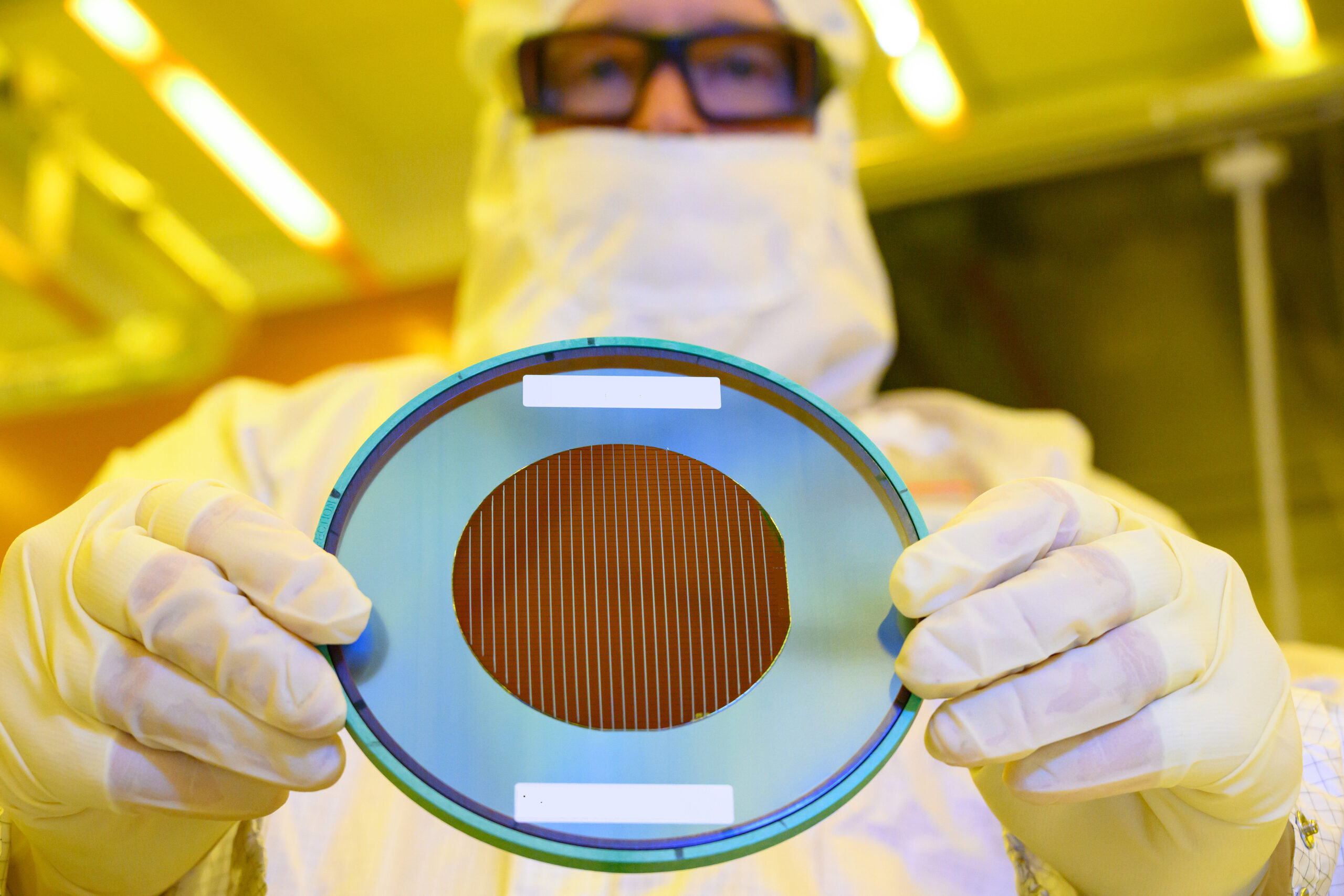 A person in a cleanroom suit and gloves holds up a silicon wafer in a laboratory. The wafer has a reflective surface with intricate patterns. The background is softly lit in yellow tones.