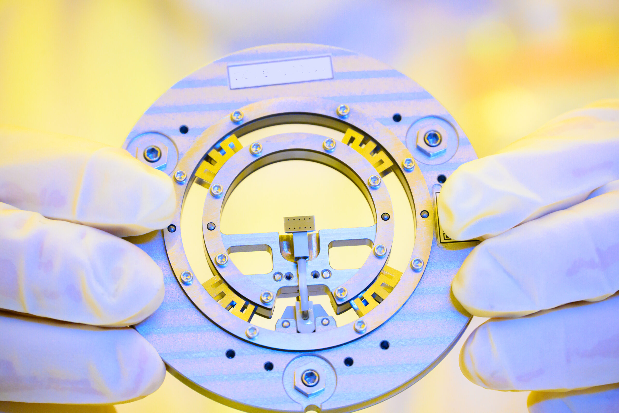 Gloved hands holding a circular metallic component with intricate internal structures, possibly a part of a mechanical or electronic device, against a blurred yellow background.