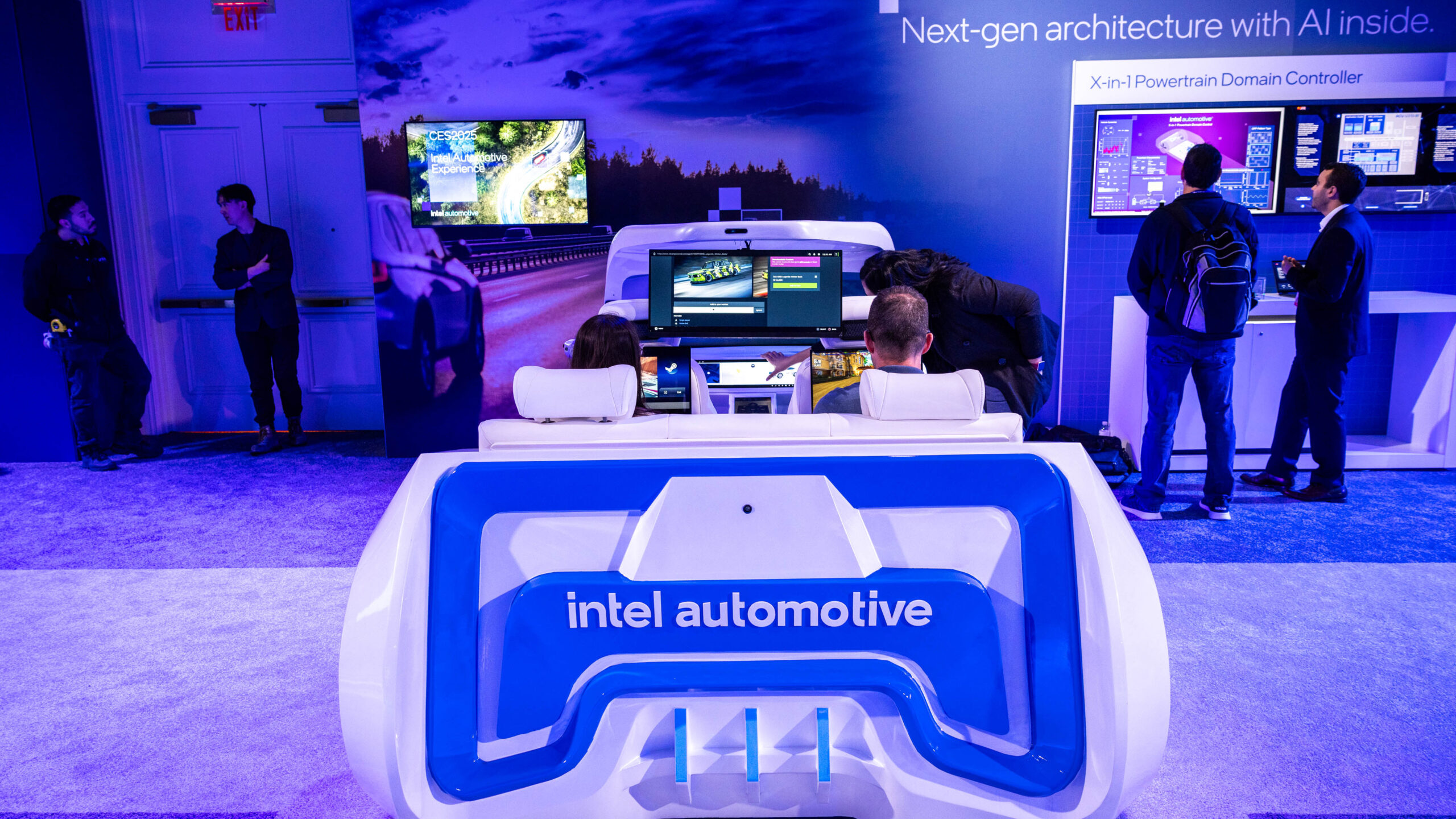People are interacting with displays and a mock car setup at an Intel Automotive exhibit. The booth highlights next-gen architecture with AI inside. The scene is lit with blue and purple tones, creating a tech-focused atmosphere.