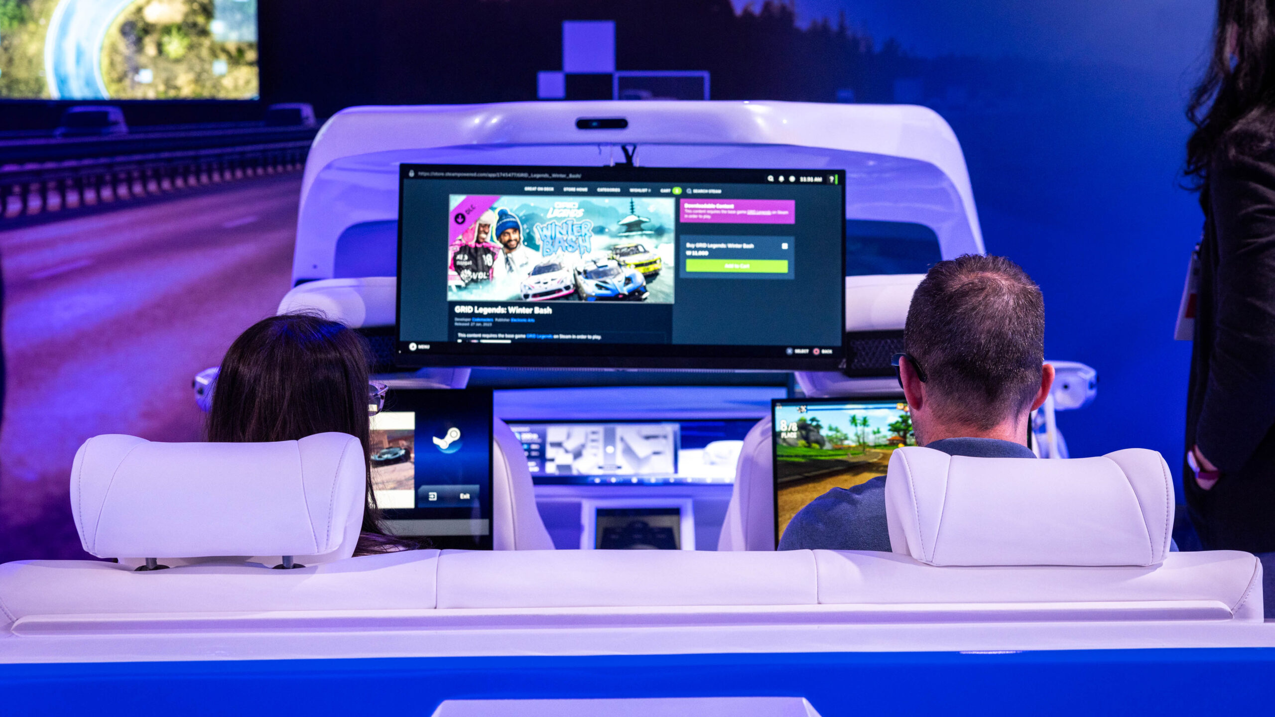 Two people sit in white car seats facing a large digital screen displaying a video game interface. Each person holds a tablet. The setting resembles a high-tech car showroom with a blue ambient light.