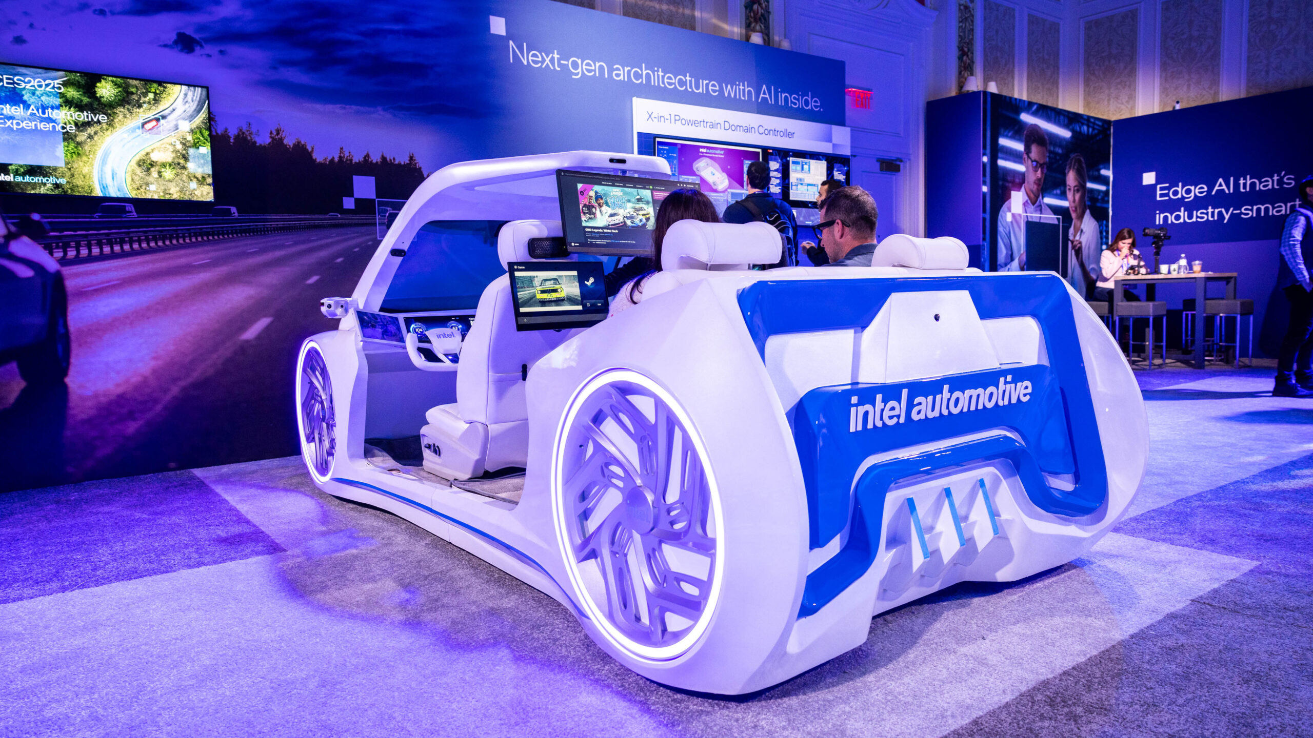 A futuristic, open-concept car with Intel Automotive branding is displayed at a tech expo. The interior features screens and advanced controls. Attendees are seated inside, and informational boards about AI technology are visible in the background.