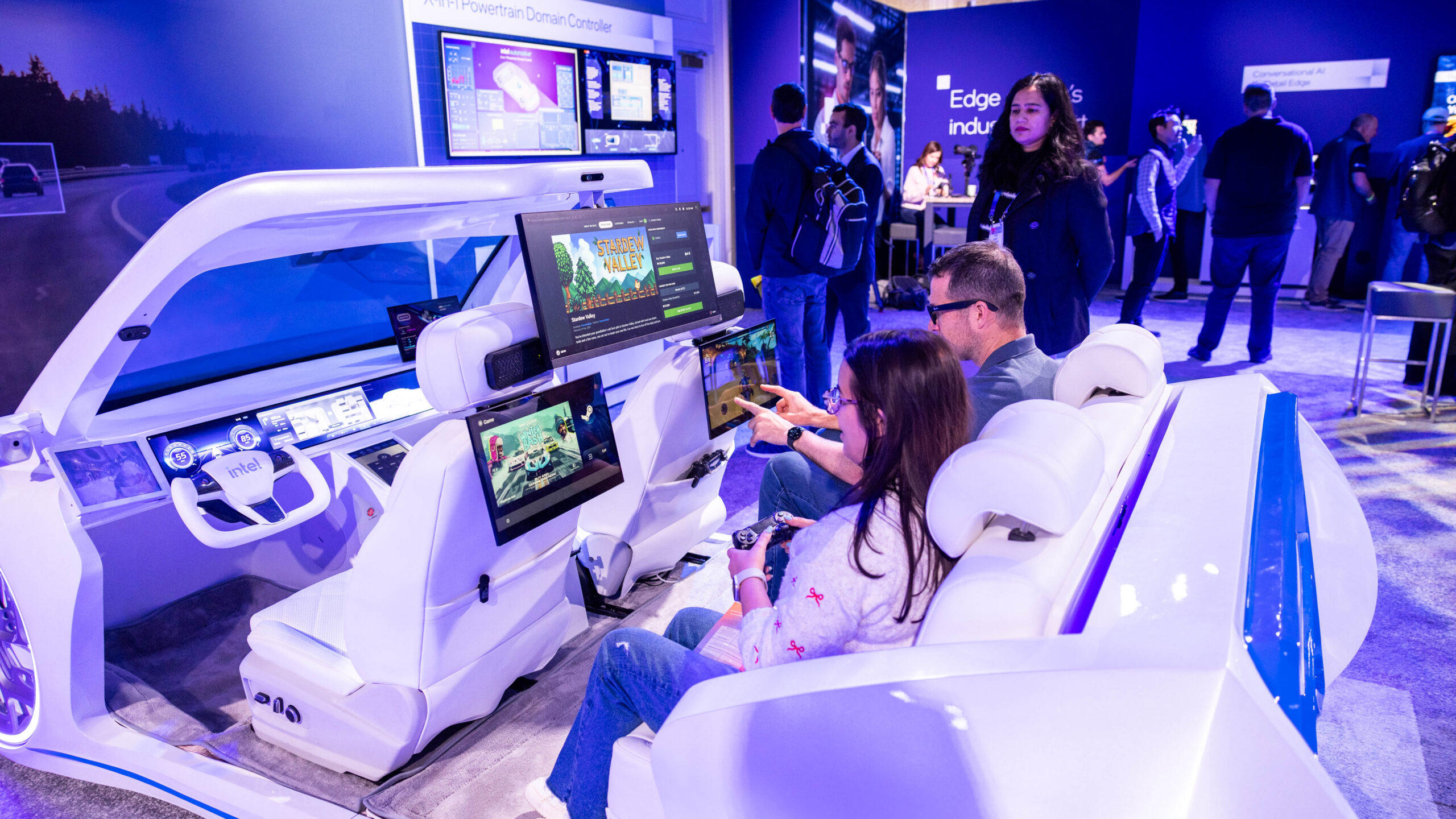 People interacting with a futuristic car interior exhibit, featuring large screens and advanced technology, at a tech event. The area has a purple-lit ambiance, and several individuals are visible in the background.
