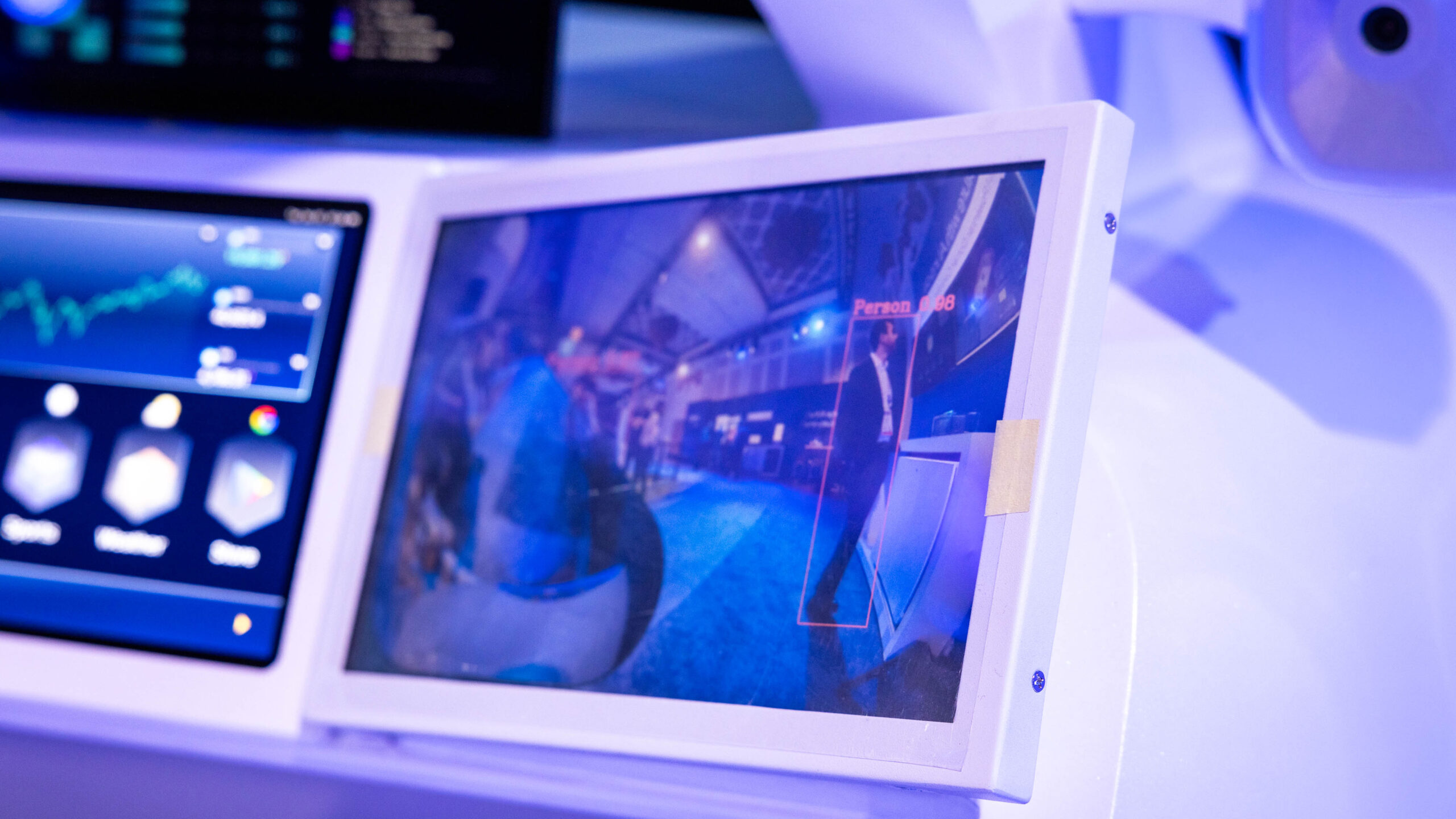 A tablet displaying a security camera feed with a highlighted figure and the label Person. Adjacent screens show graphic data in a bright, modern room with blue lighting.