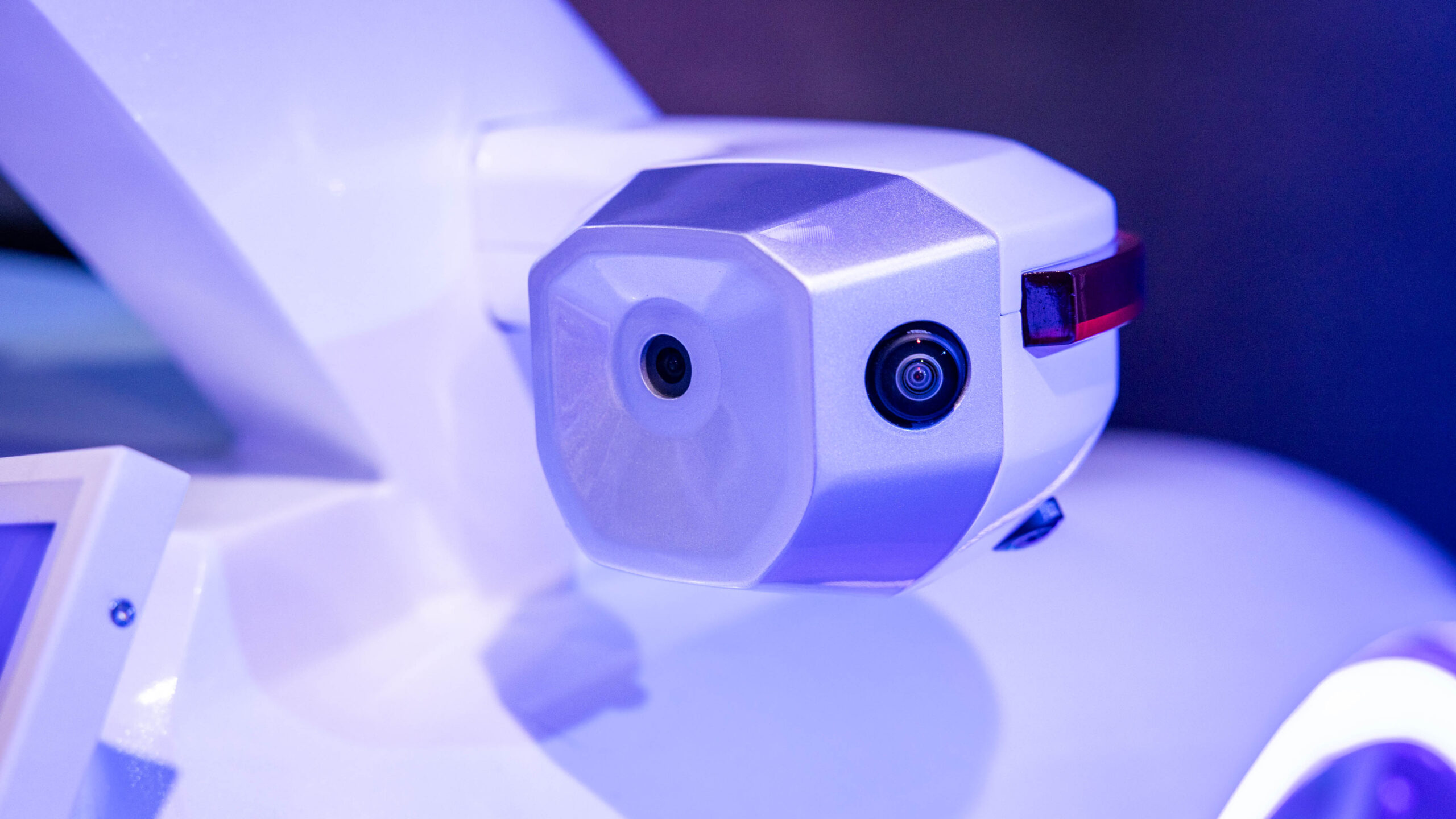Close-up of a white robotic camera with two lenses, possibly used for surveillance or monitoring. The device is mounted on a smooth surface, surrounded by a blue-toned background, suggesting a technological or futuristic setting.