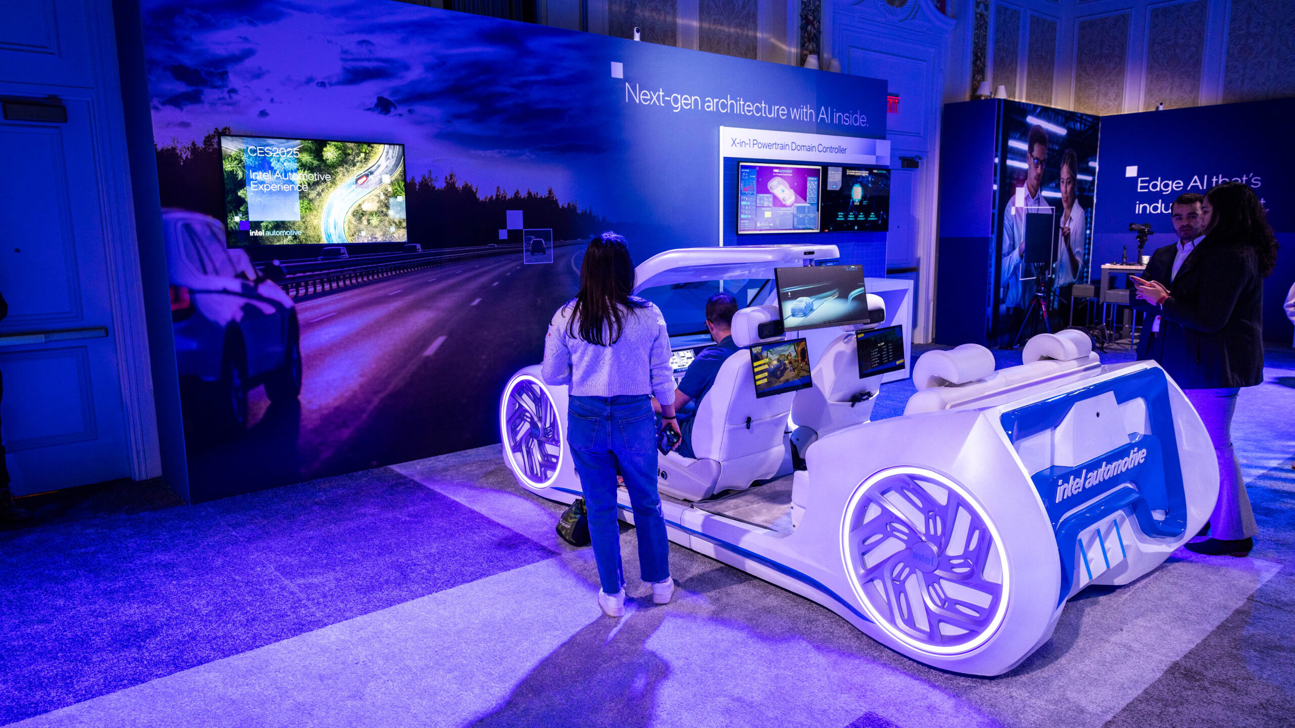 People interact with a futuristic car exhibit featuring large screens and advanced technology displays. The exhibit is set in a dimly lit room with blue and purple lighting, showcasing a Next-gen architecture theme on the wall.
