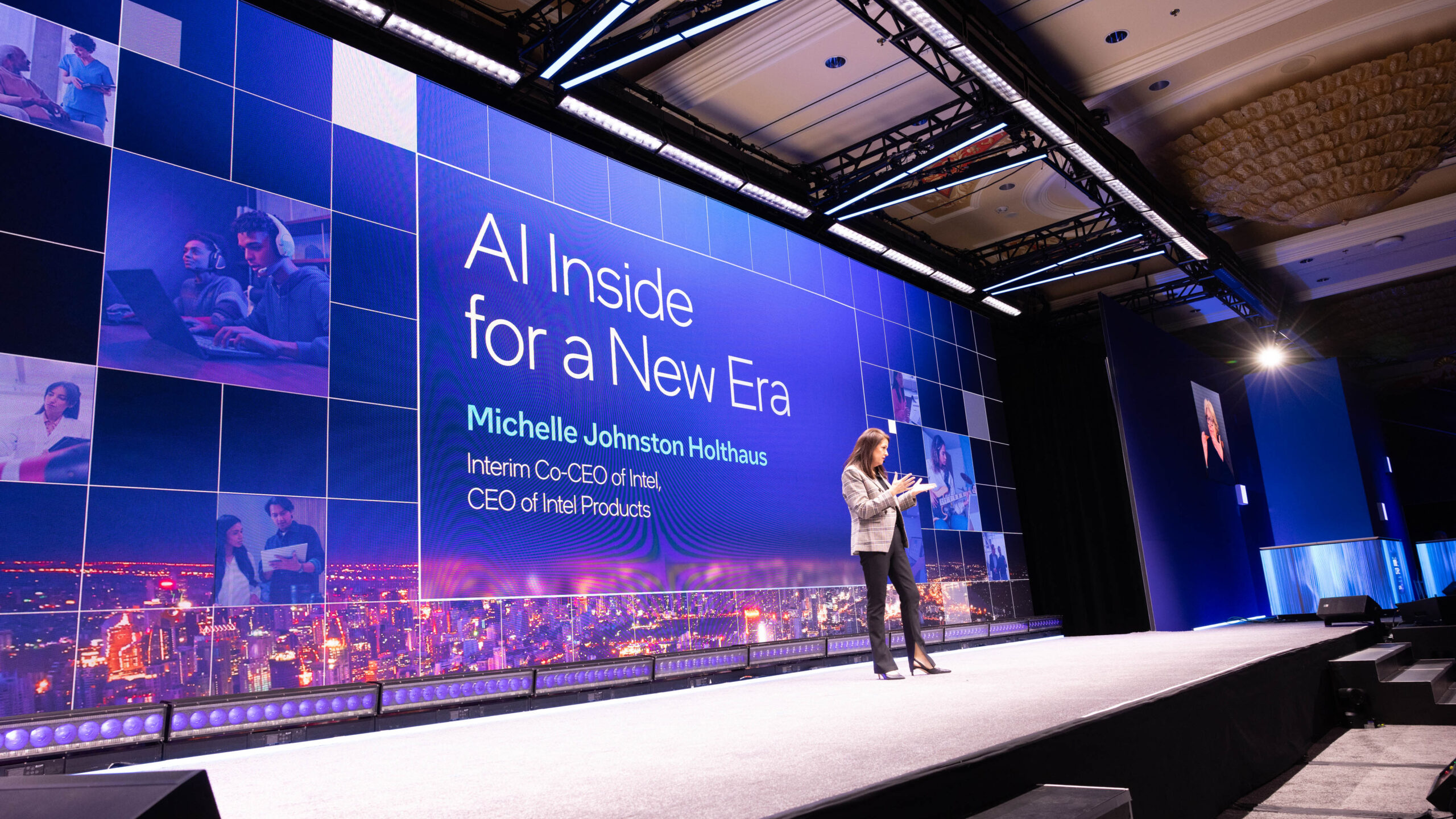 A speaker stands on a stage in front of a large screen displaying the text AI Inside for a New Era and Michelle Johnston Holthaus, Interim Co-CEO of Intel, CEO of Intel Products. The stage is lit with blue and purple lights.