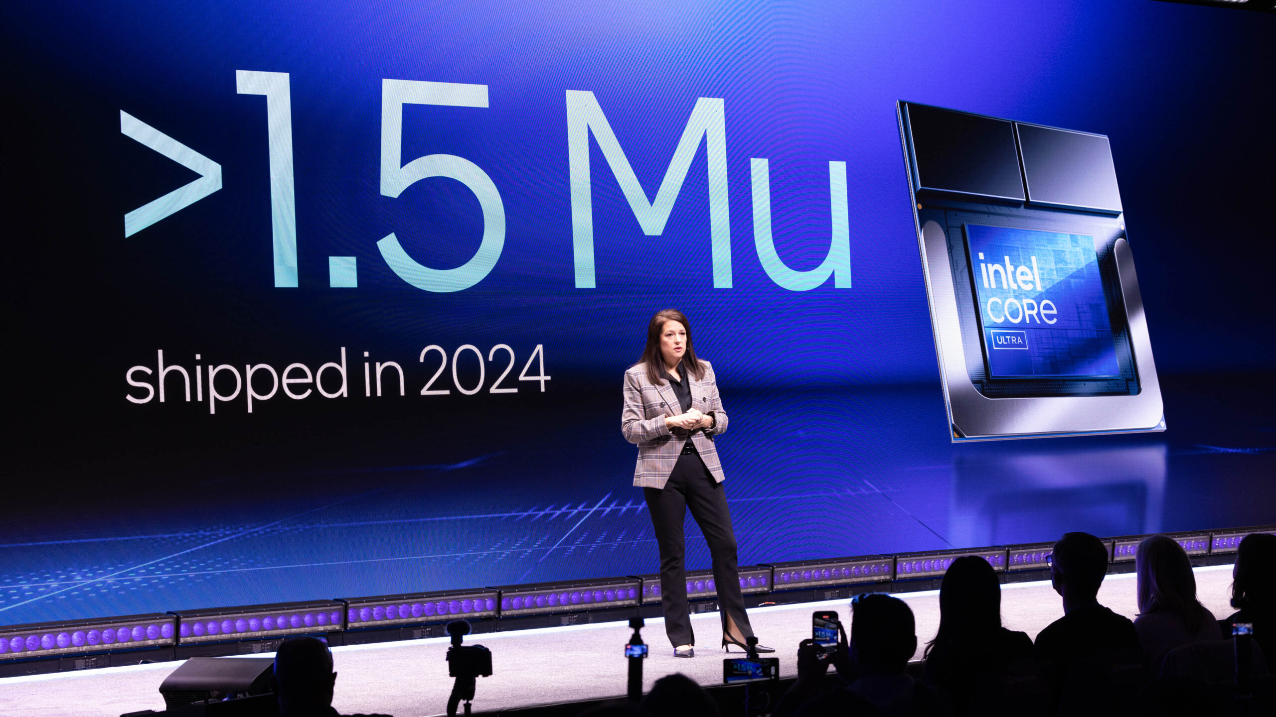 A presenter stands on a stage with a large screen displaying >1.5 Mu shipped in 2024 and an image of an Intel Core Ultra chip. The audience is visible in silhouette.