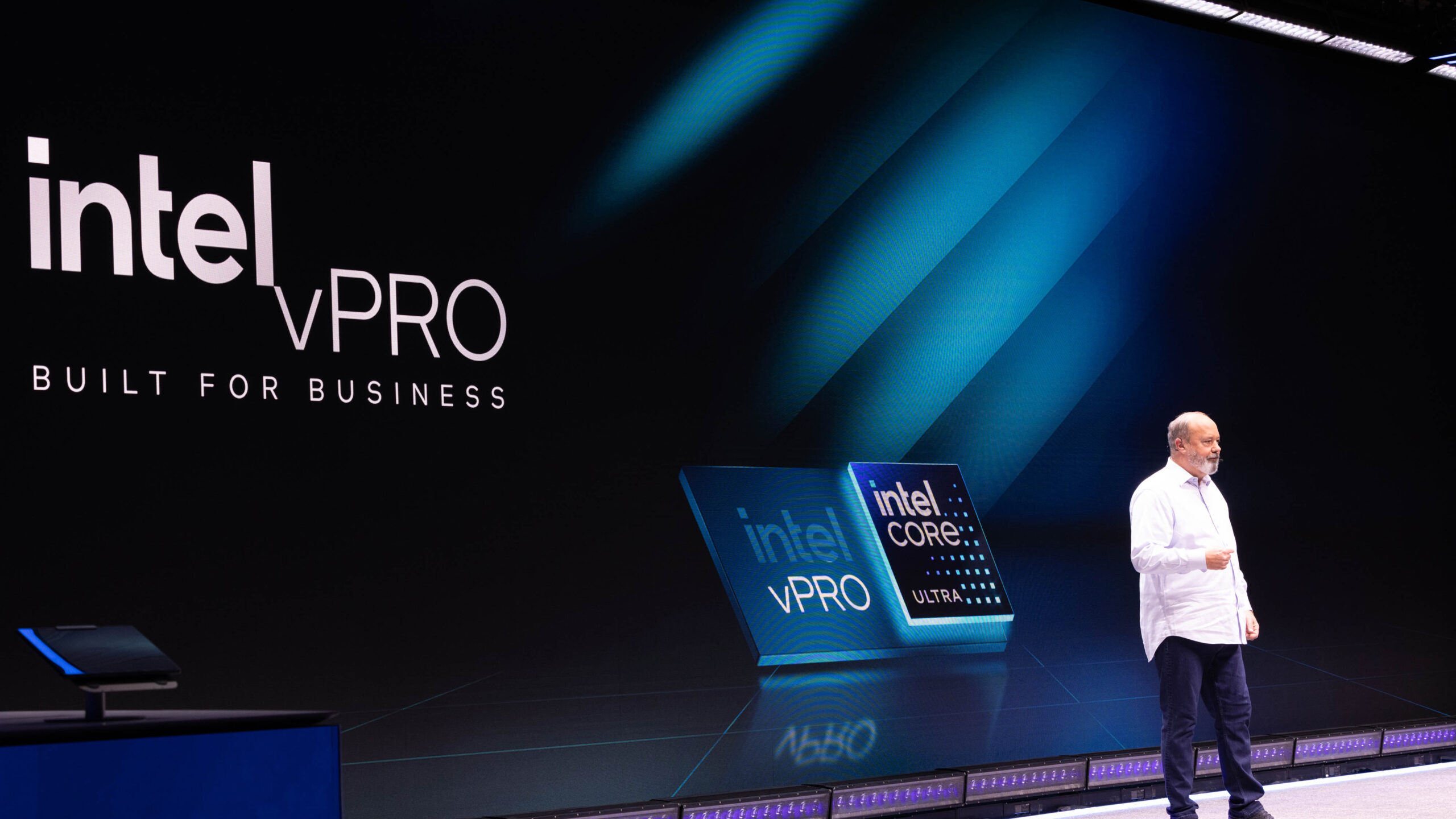 A person stands on a stage giving a presentation about Intel vPro, with large screens displaying the Intel vPro logo and Built for Business” slogan. A digital display features the Intel Core and vPro logos.
