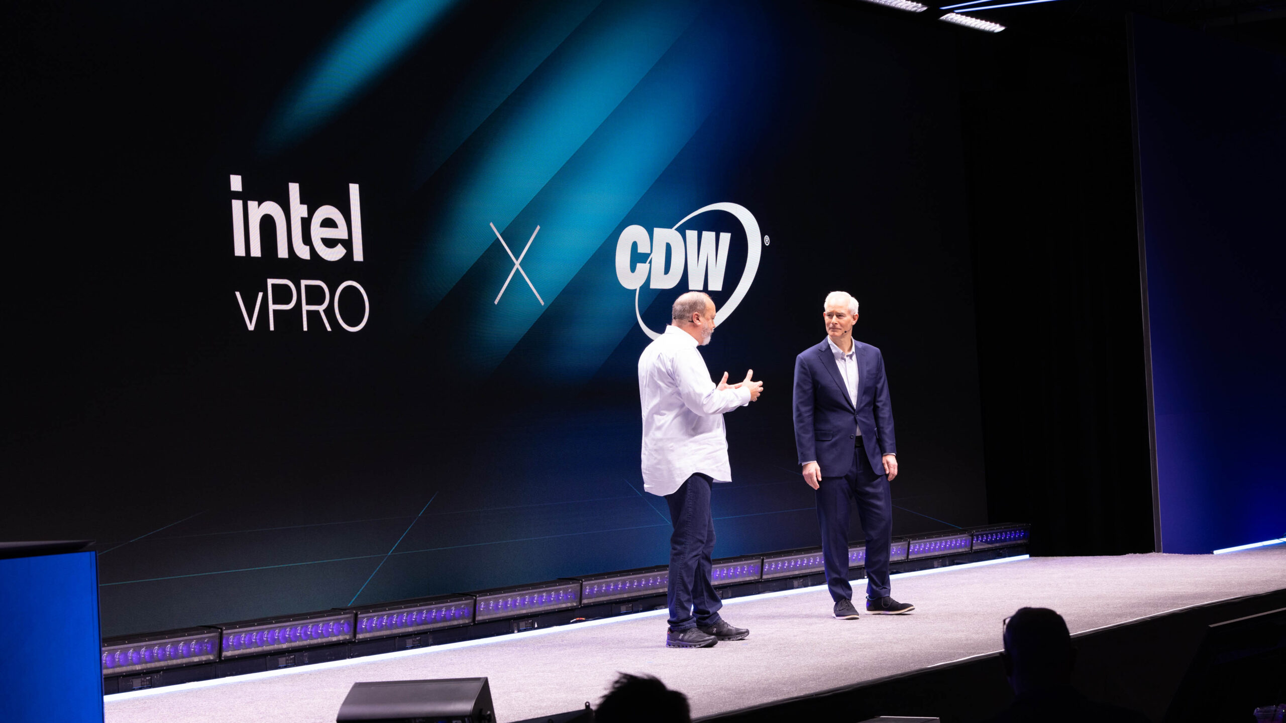Two men stand on a stage in front of a large screen displaying the Intel vPro and CDW logos. One man gestures while speaking, and both are wearing suits. The stage is lit with spotlights, and the background features a blue gradient design.