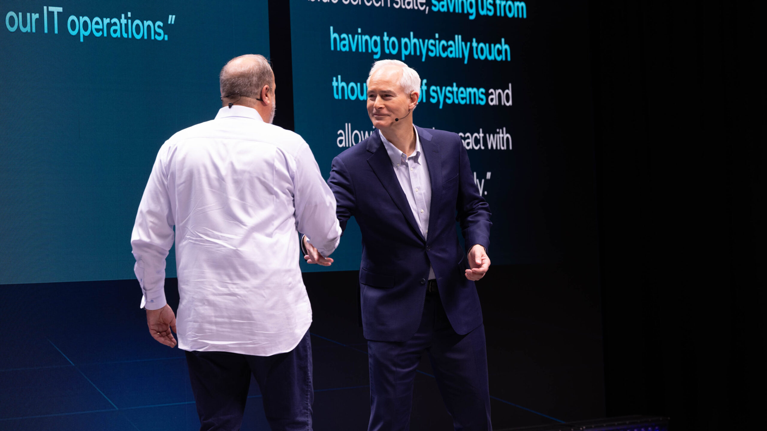 Two men are shaking hands on a stage with text displayed on large screens behind them. One man is facing the audience wearing a dark suit, and the other is wearing a white shirt and jeans. The setting appears to be a formal presentation or conference.